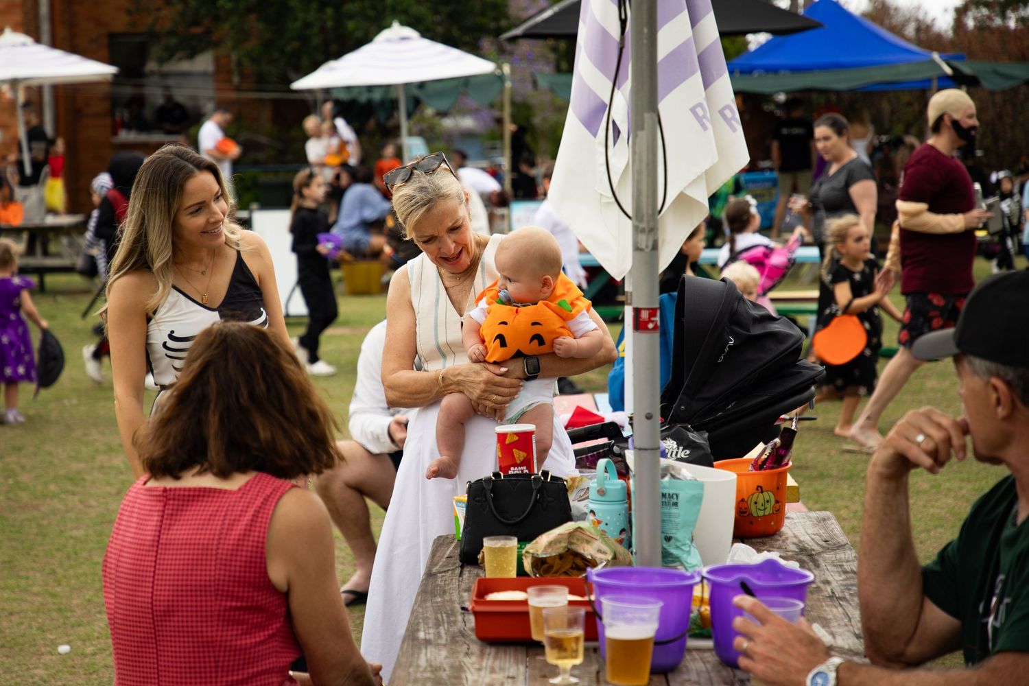 A woman holds a baby dressed in an orange Halloween costume at a festive outdoor gathering.
