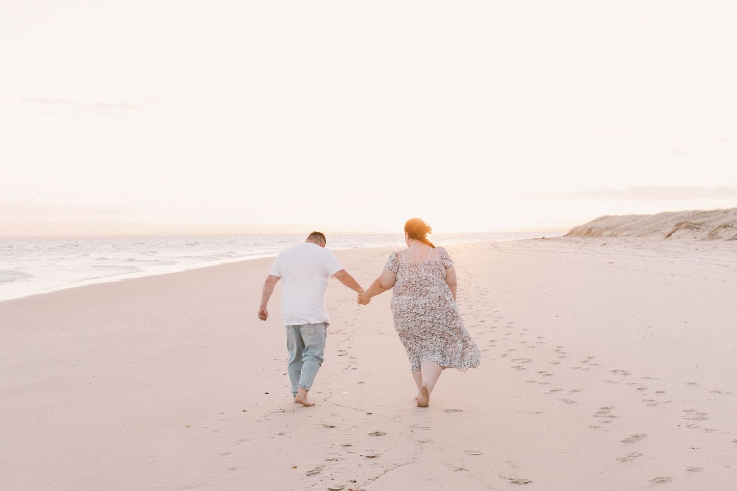 Couple walking hand in hand on sandy beach at sunset leaving footprints in the sand.