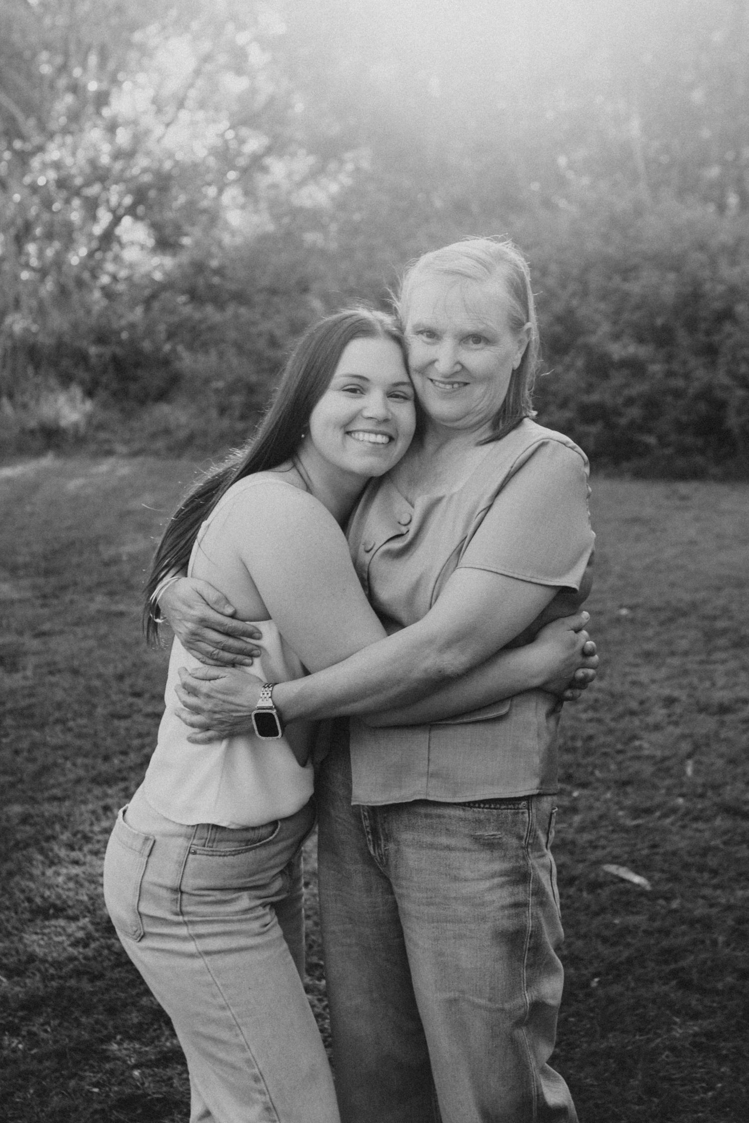 Black and white photo of two women hugging and smiling outdoors with trees in the background.