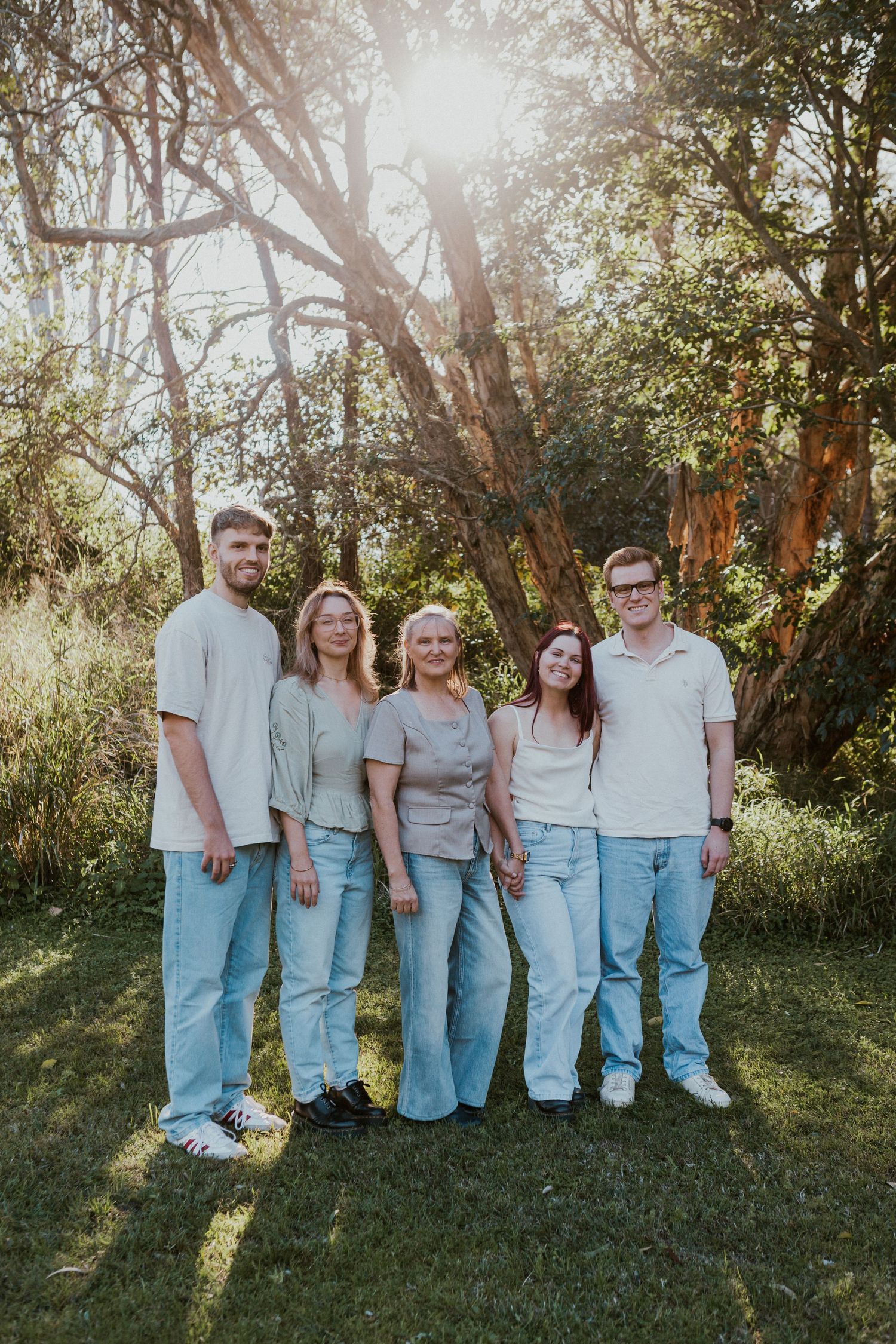 Family of five smiling outdoors in coordinated neutral outfits, standing on grass with sunlit trees in background.
