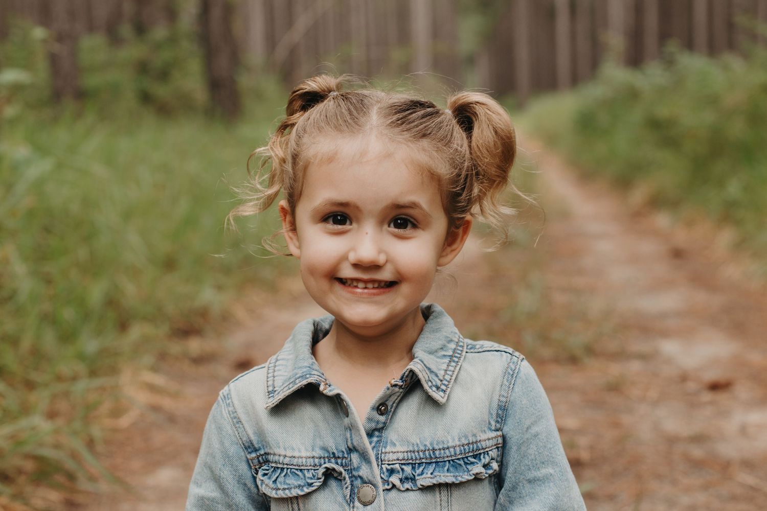 A cute smiling young girl with pigtails in a denim jacket stands on a forest dirt path.