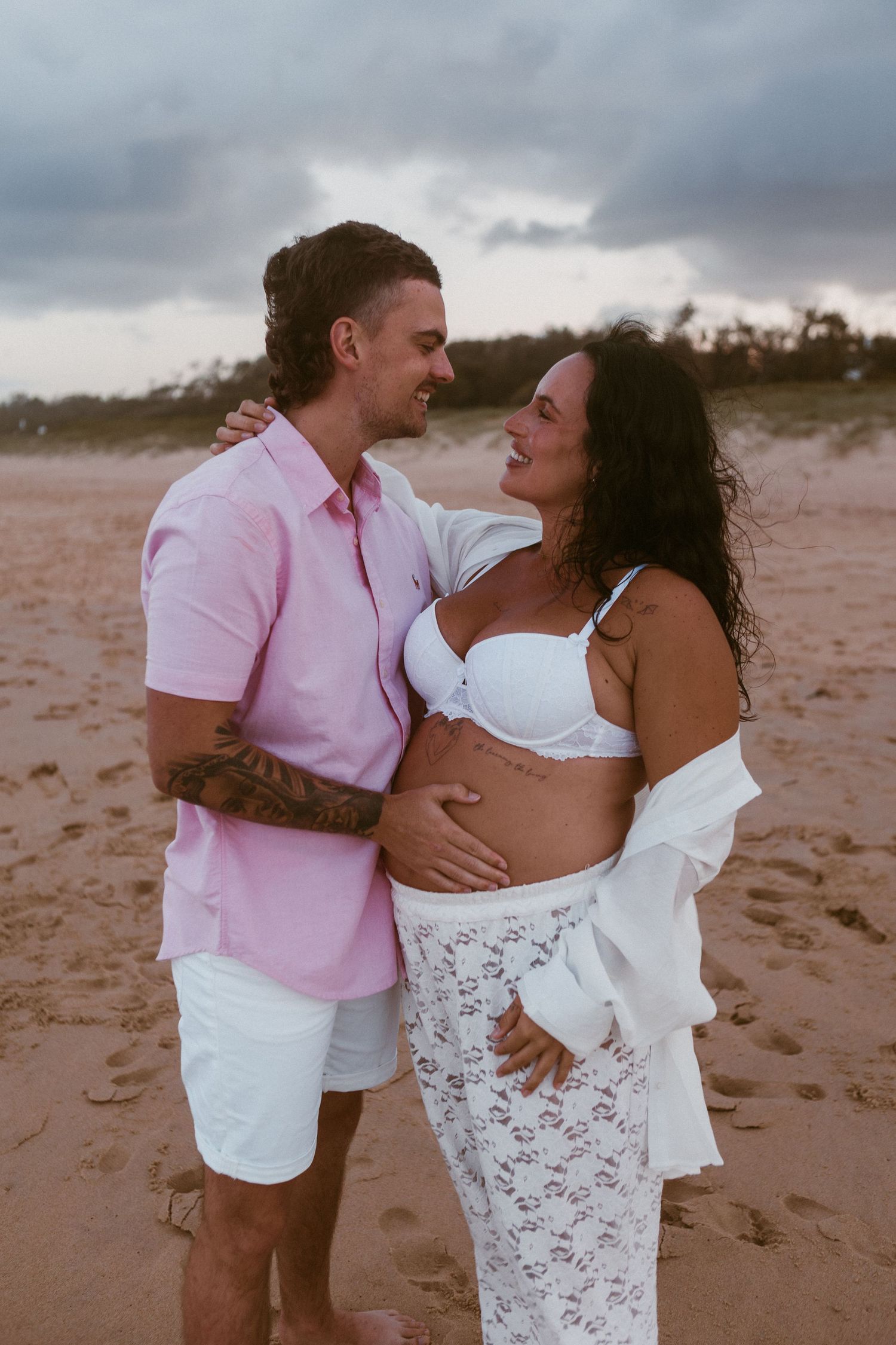 A couple shares a tender moment on a sandy beach, the woman visibly pregnant, dressed in white lace, as her partner embraces her.