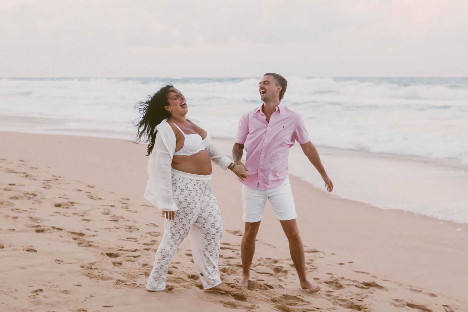 Pregnant couple holding hands and walking on a sandy beach near ocean waves during a maternity photoshoot.