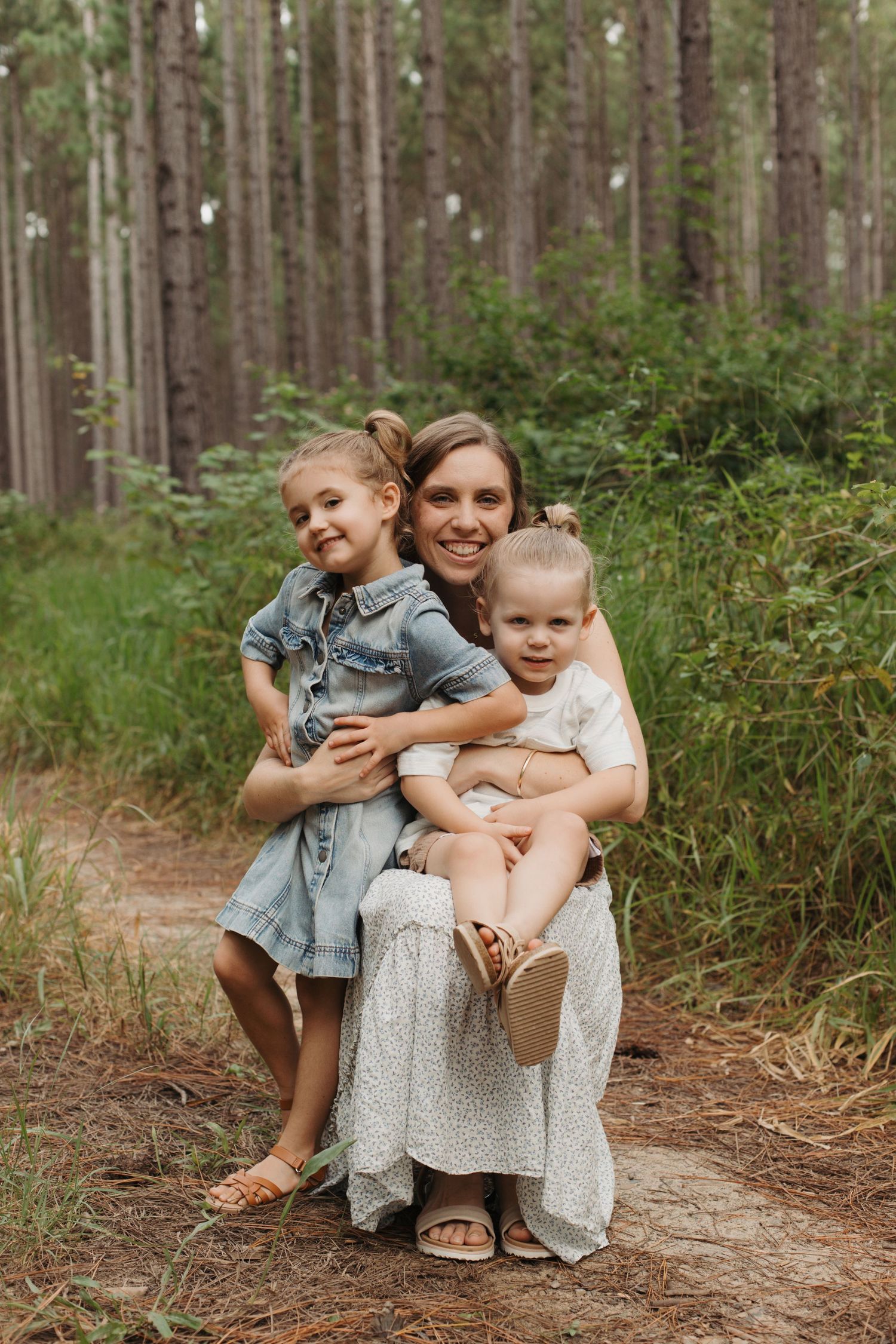 Happy mother sits on forest path hugging her two young daughters among pine trees.