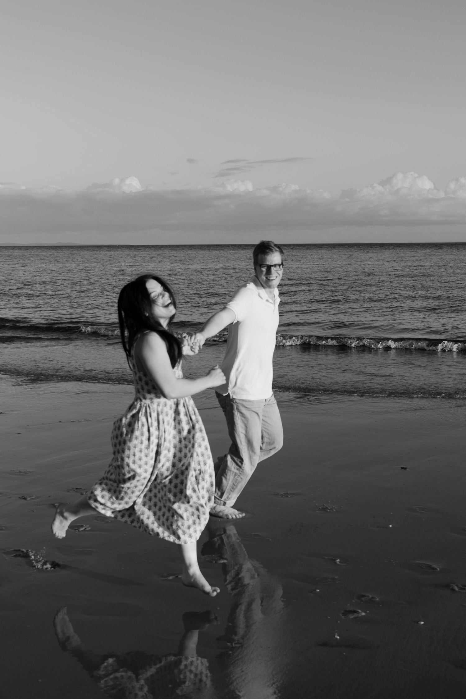 Black and white photo of a couple holding hands and dancing playfully on a beach at sunset.