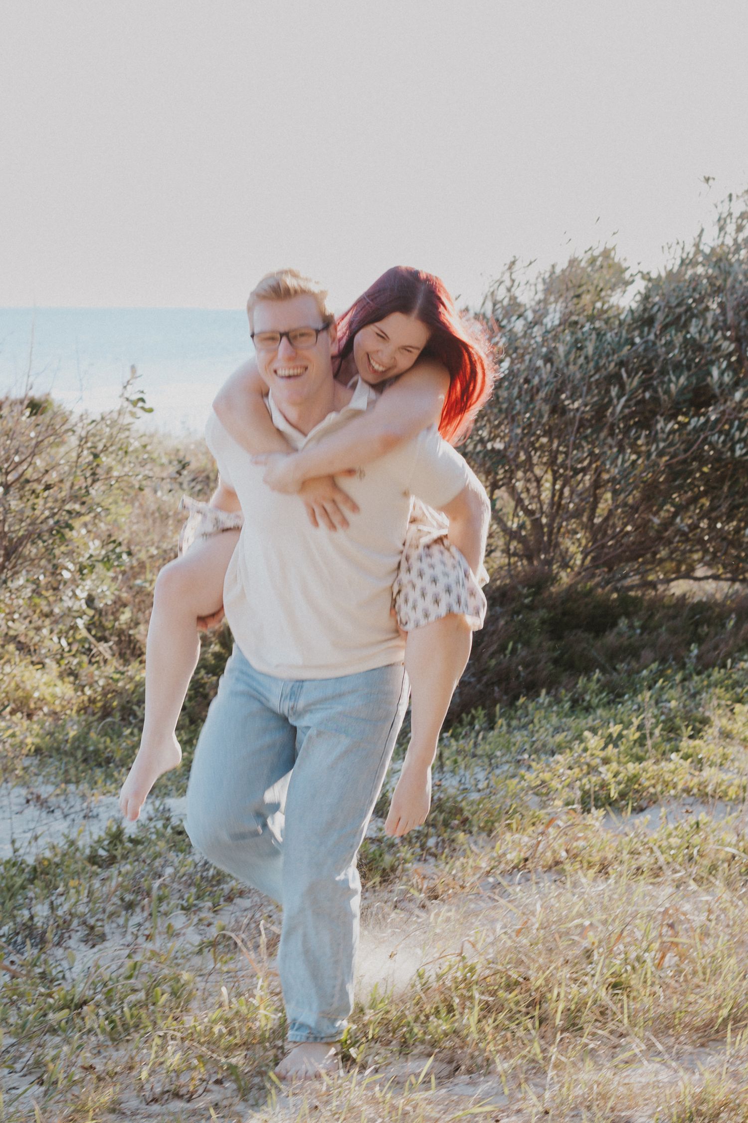A happy couple laughs as man gives woman a piggyback ride near coastal dunes.