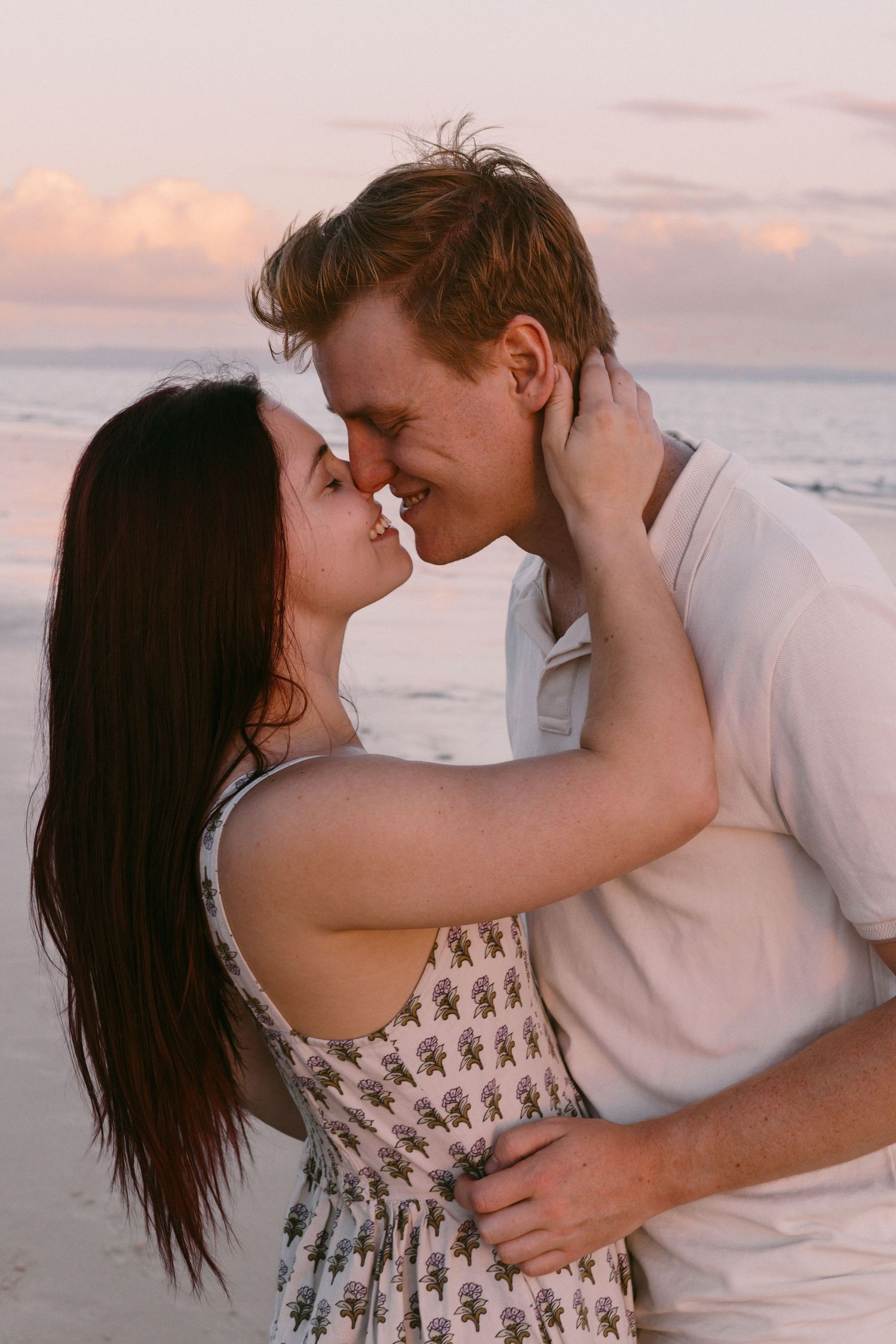 Couple sharing a romantic nose-to-nose moment on a beach at sunset with ocean backdrop.