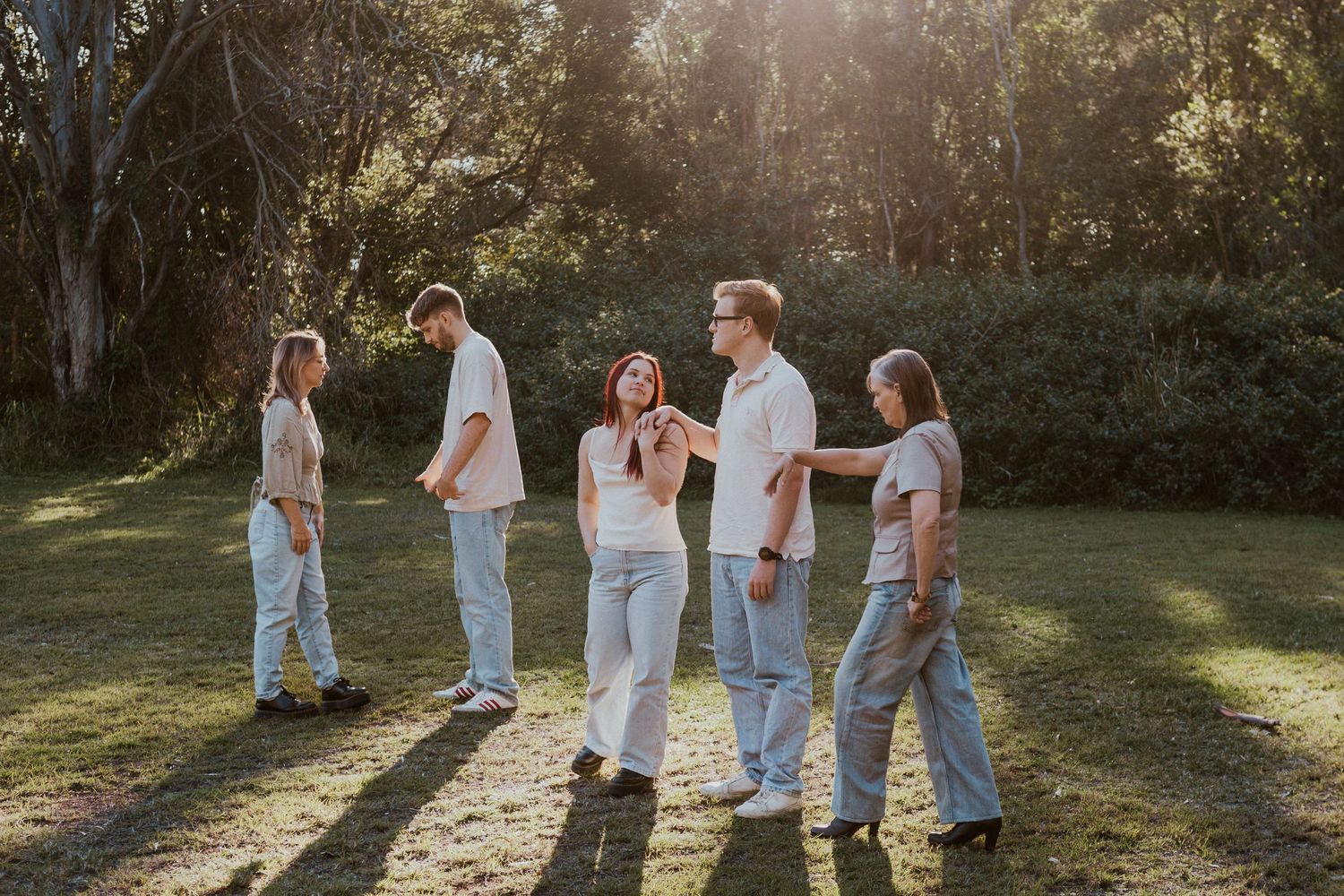 Five young adults in casual denim outfits laughing and interacting outdoors in a sunlit park surrounded by trees.
