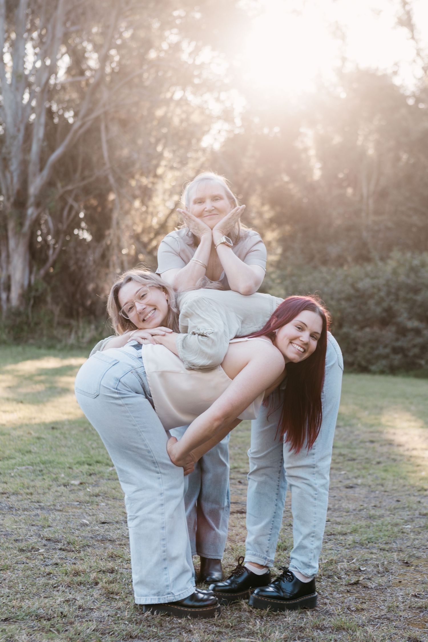 Three women posing playfully outdoors in a sunlit park, smiling and stacking together for a fun group photo.