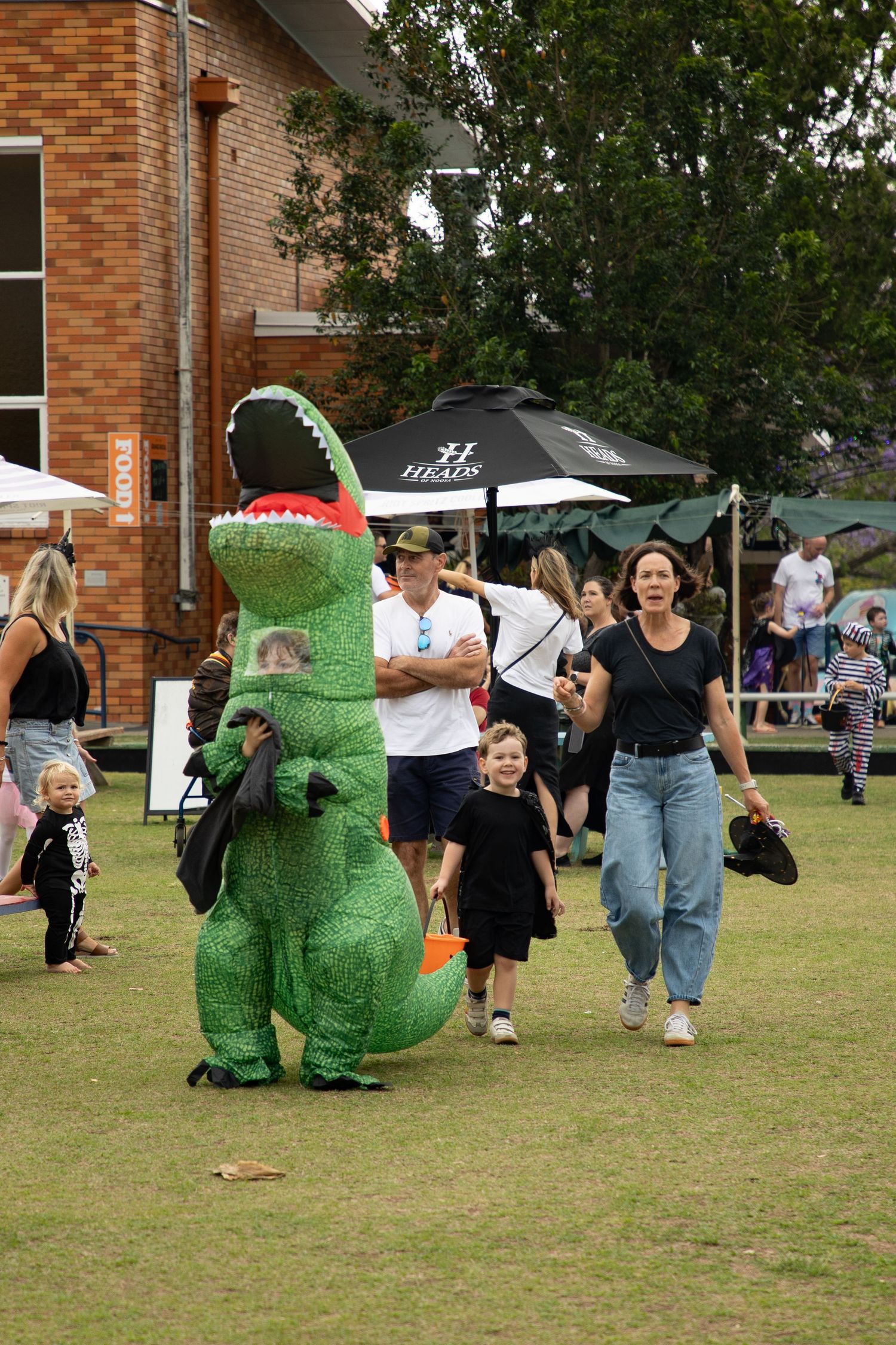 A person in an inflatable T-Rex costume walks among festival crowds at a Halloween outdoor event.