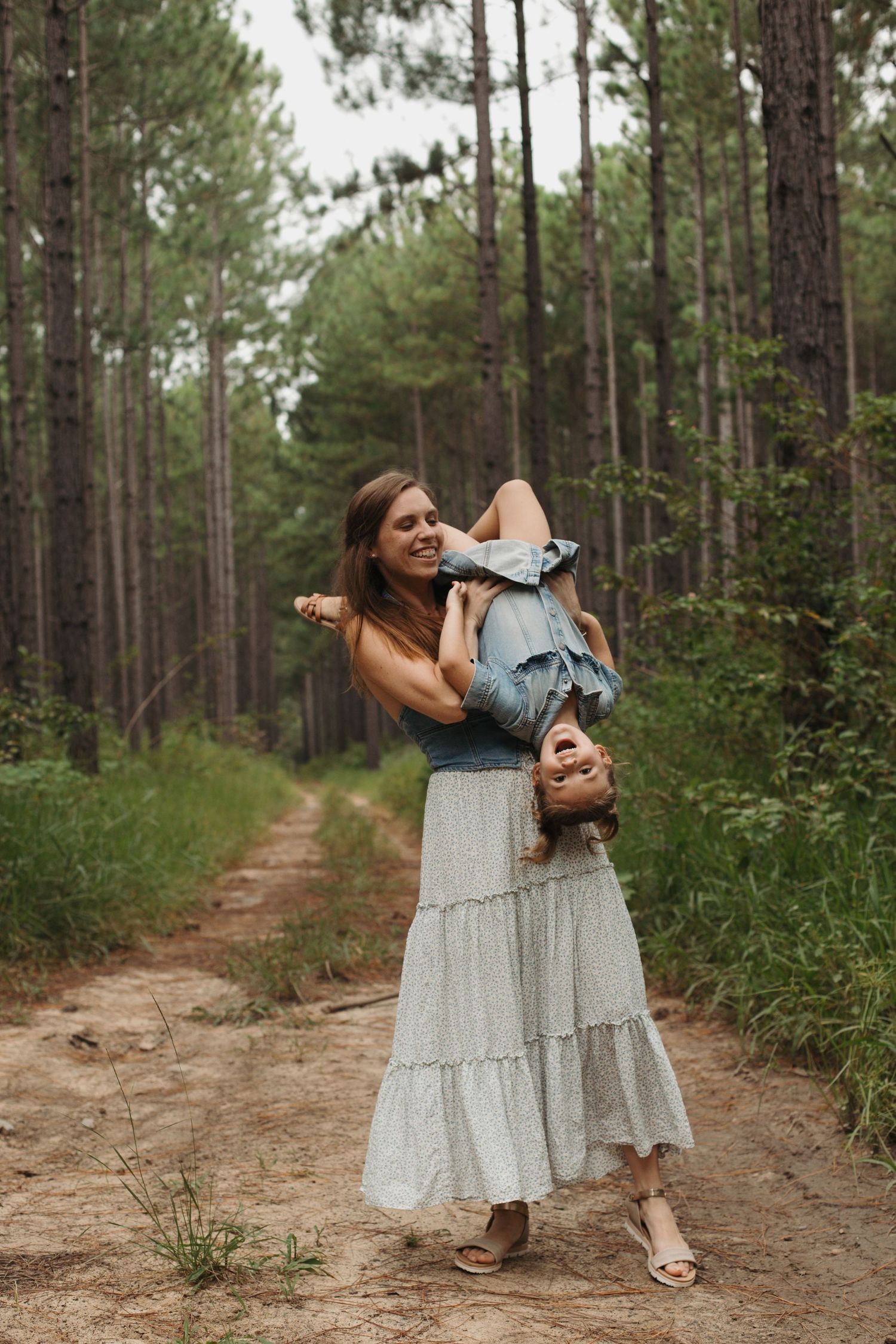 Smiling mother playfully holds child upside down on a pine forest trail.