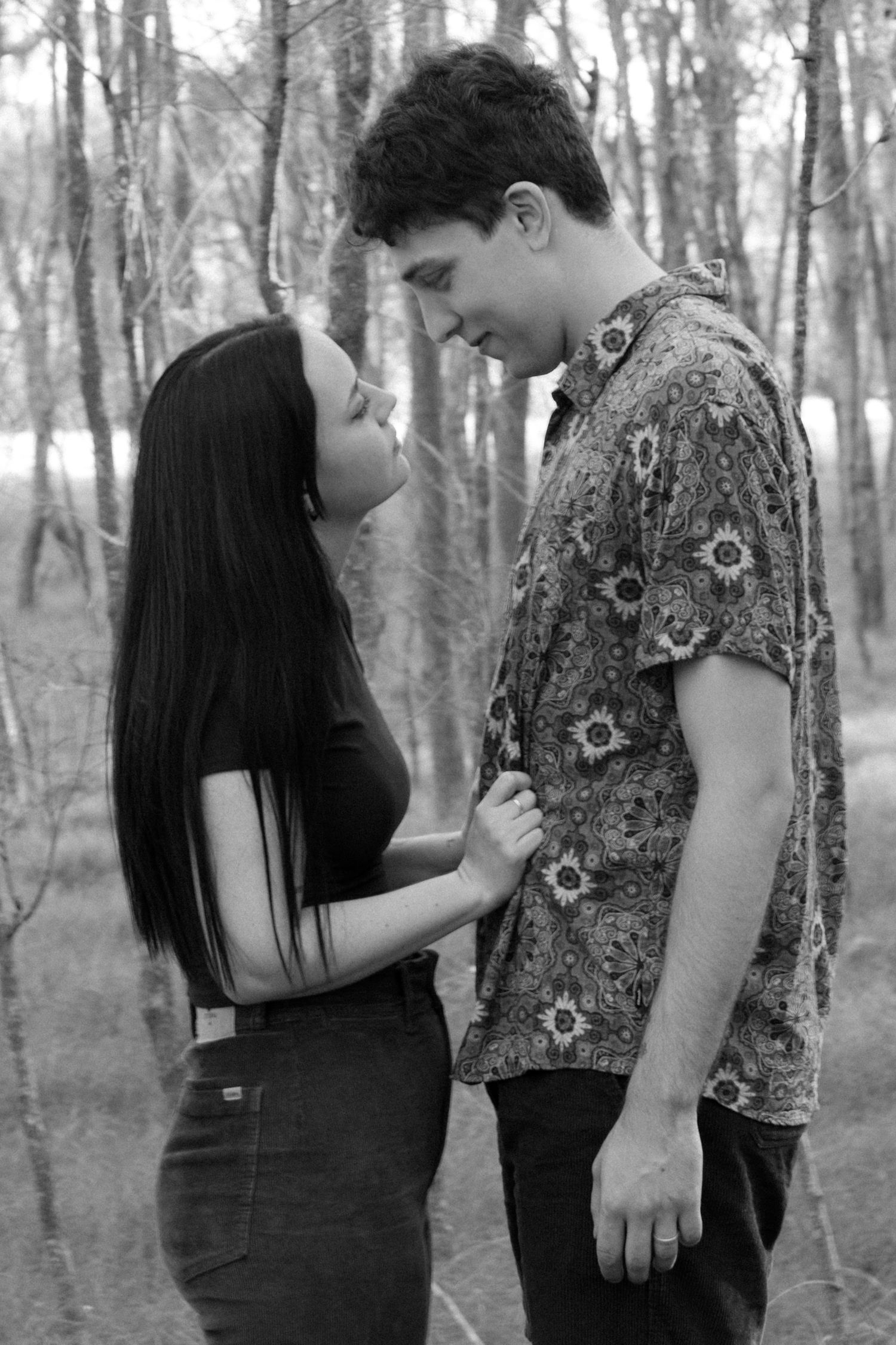 Black and white photo of a young couple facing each other tenderly in a forest setting.