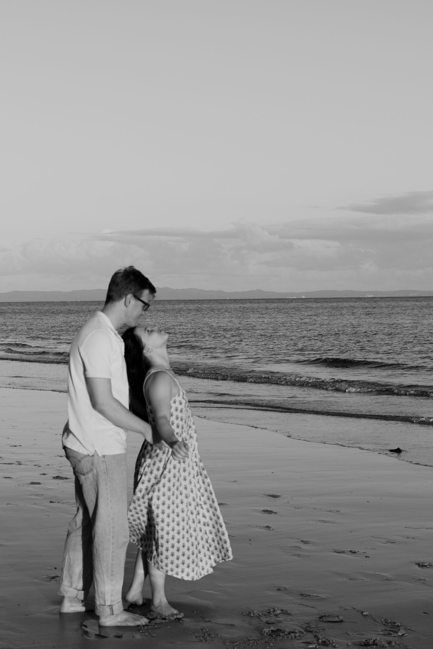Black and white photo of a romantic couple embracing on a beach at dusk by the ocean.