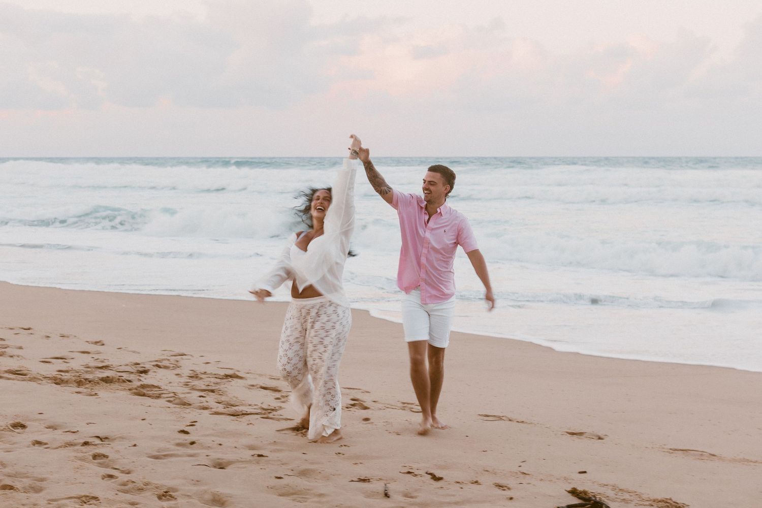 Couple dancing joyfully on a sandy beach at sunset, waves crashing in the background, wearing elegant casual attire.