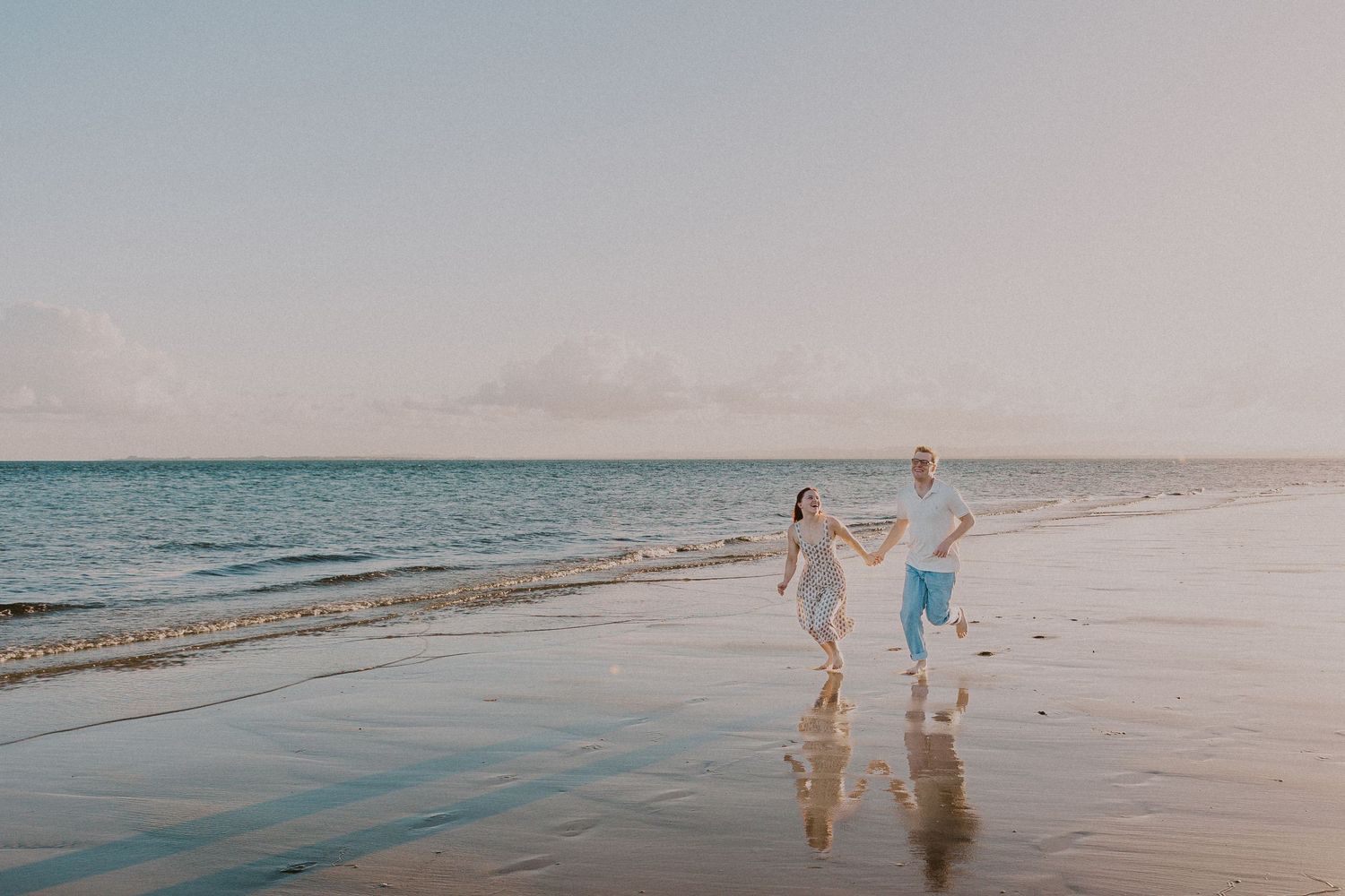Couple holding hands walking along a serene beach at sunset with calm ocean waves.