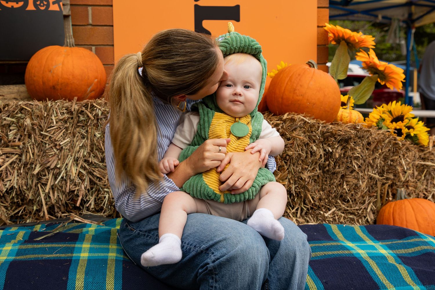 A mother kisses her baby dressed in a green costume while sitting on hay bales with pumpkins.