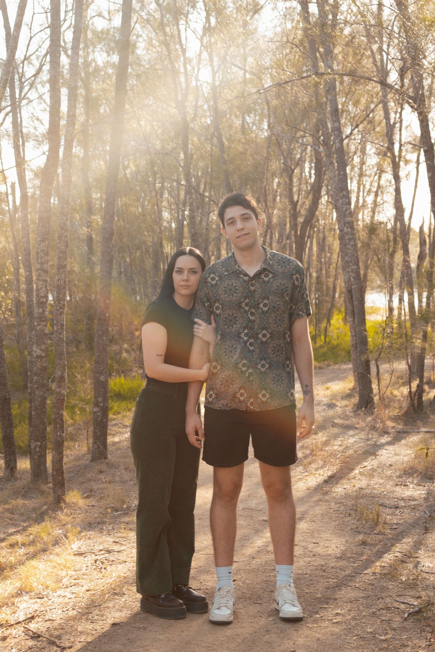 A young couple poses together on a sunlit dirt path surrounded by tall trees.