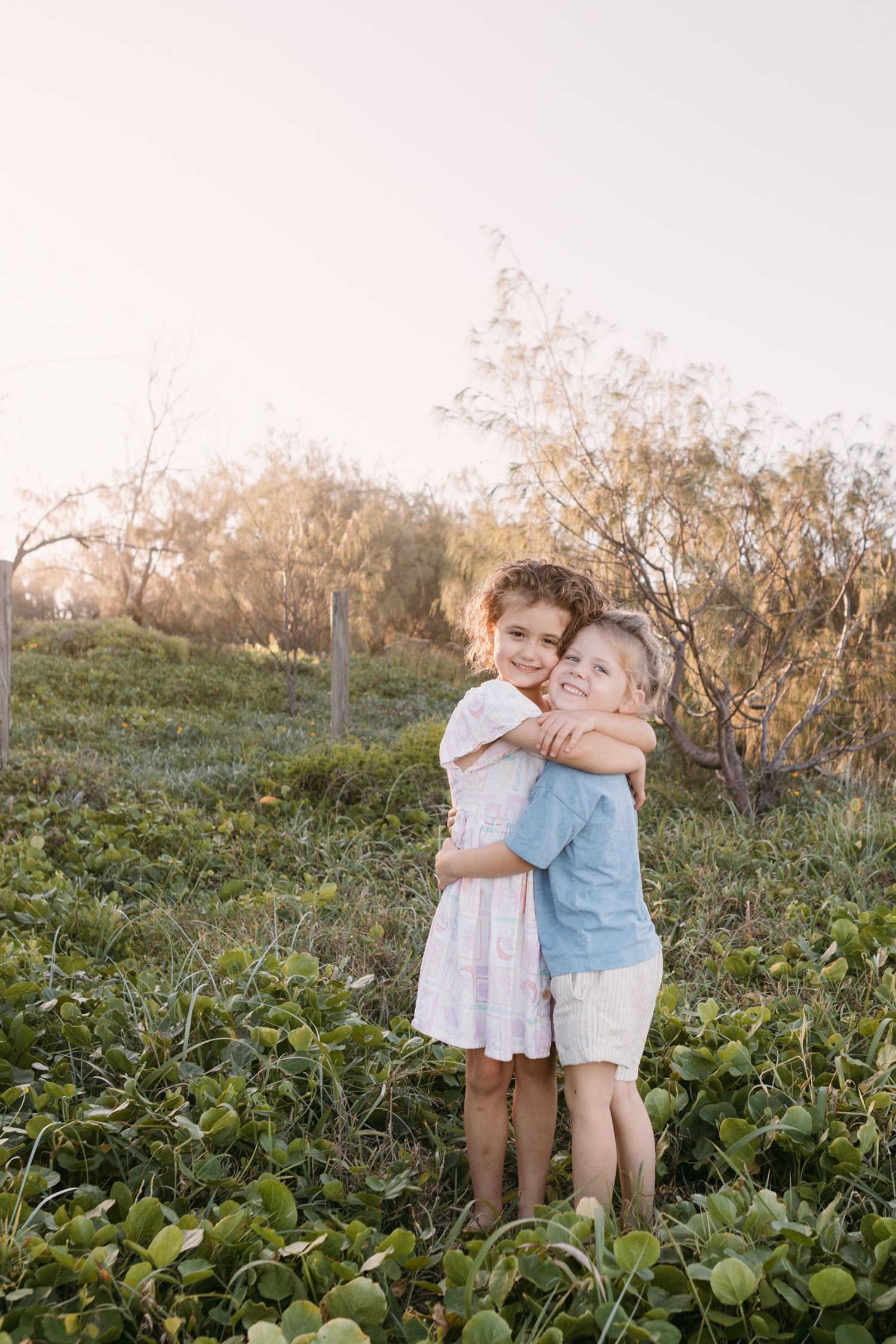 Two young children hugging warmly in a sunlit green field with trees in the background.