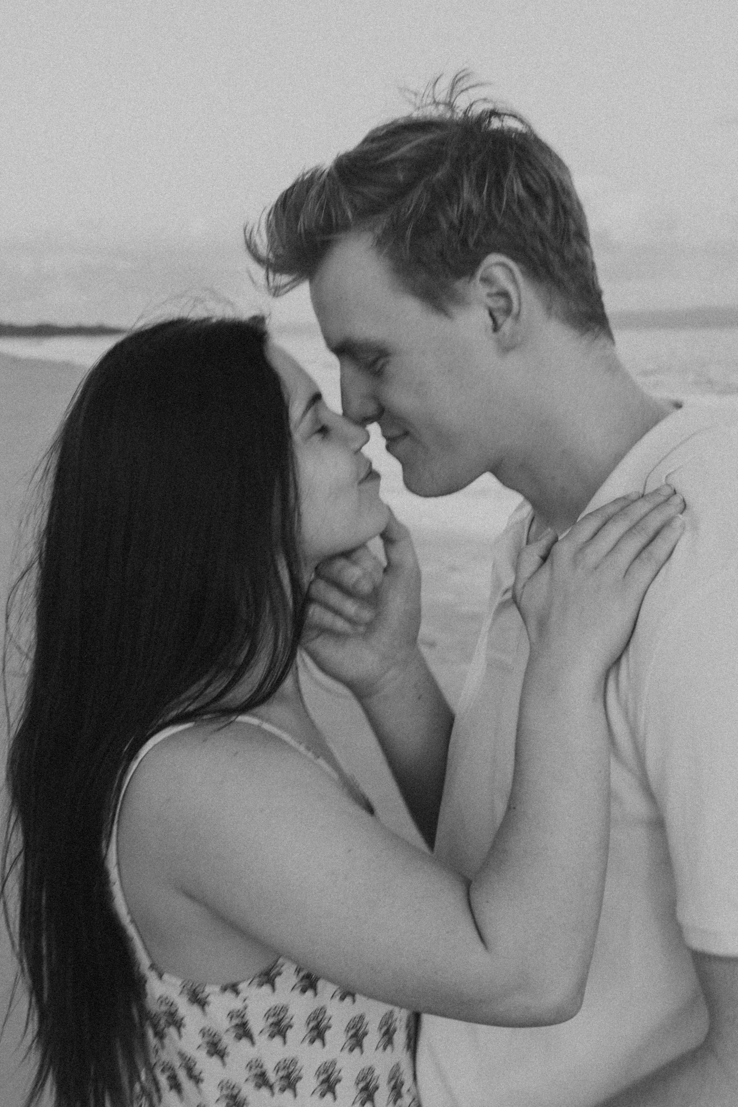 A couple shares an intimate almost-kiss moment on the beach in black and white.