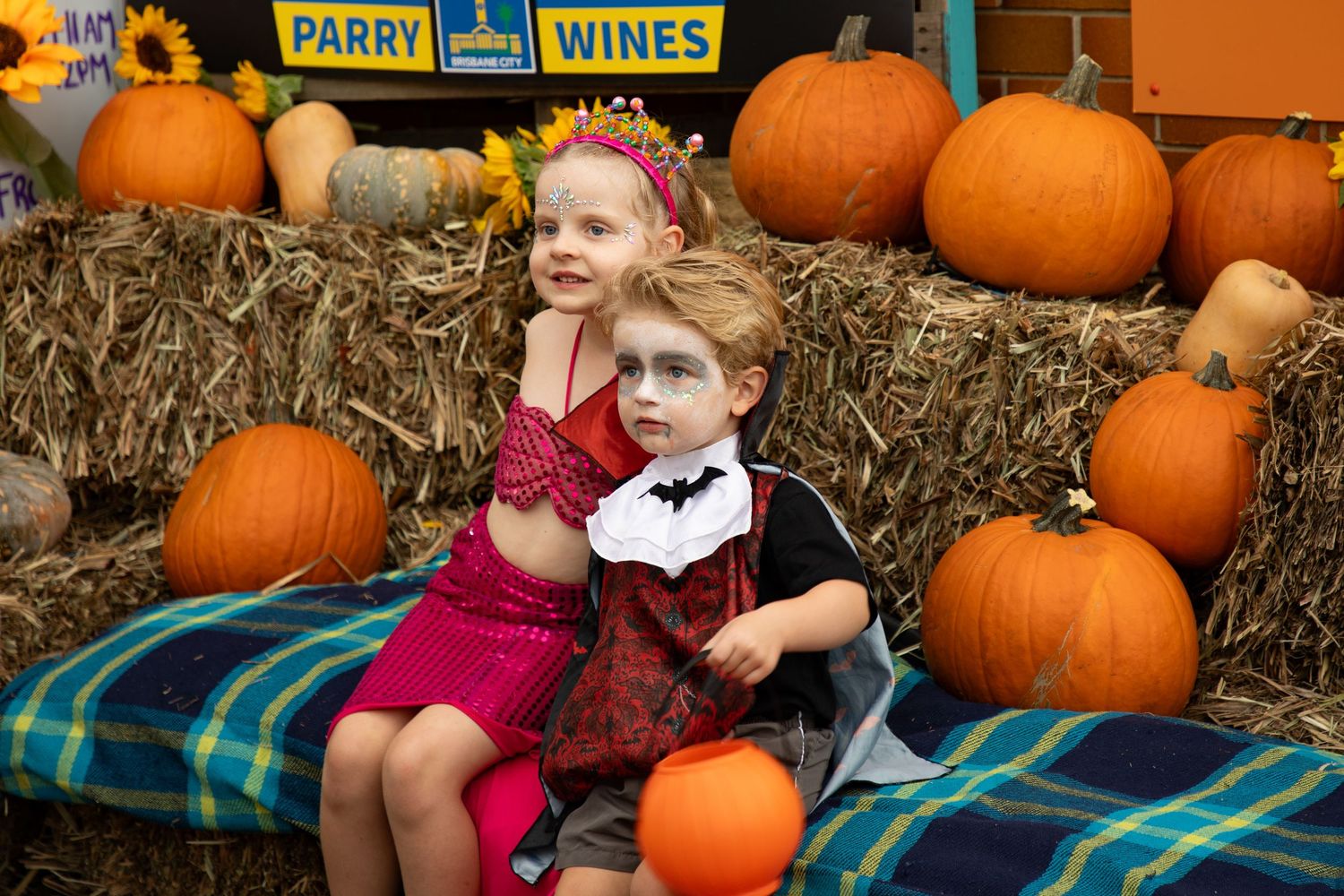 Two young children in Halloween costumes sit together surrounded by pumpkins at a festive patch.