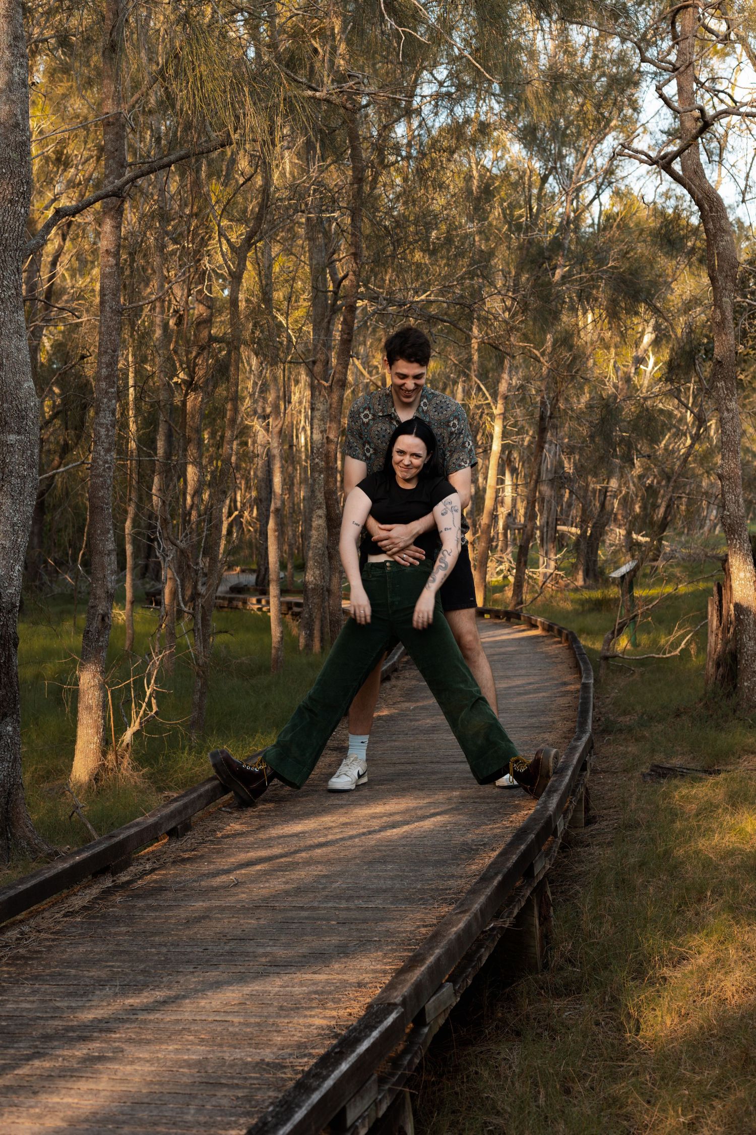 Couple laughing and playing together on a wooden boardwalk path through a sunlit eucalyptus forest.