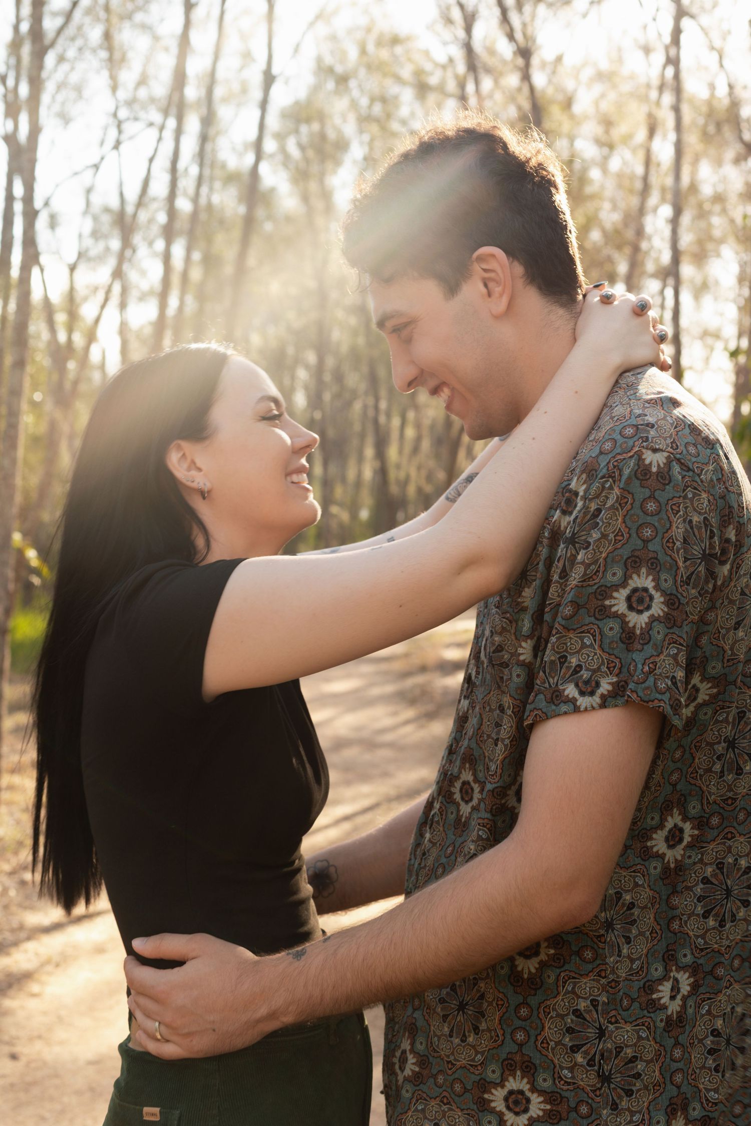 Smiling couple gazing at each other with arms around each other in sunlit forest.