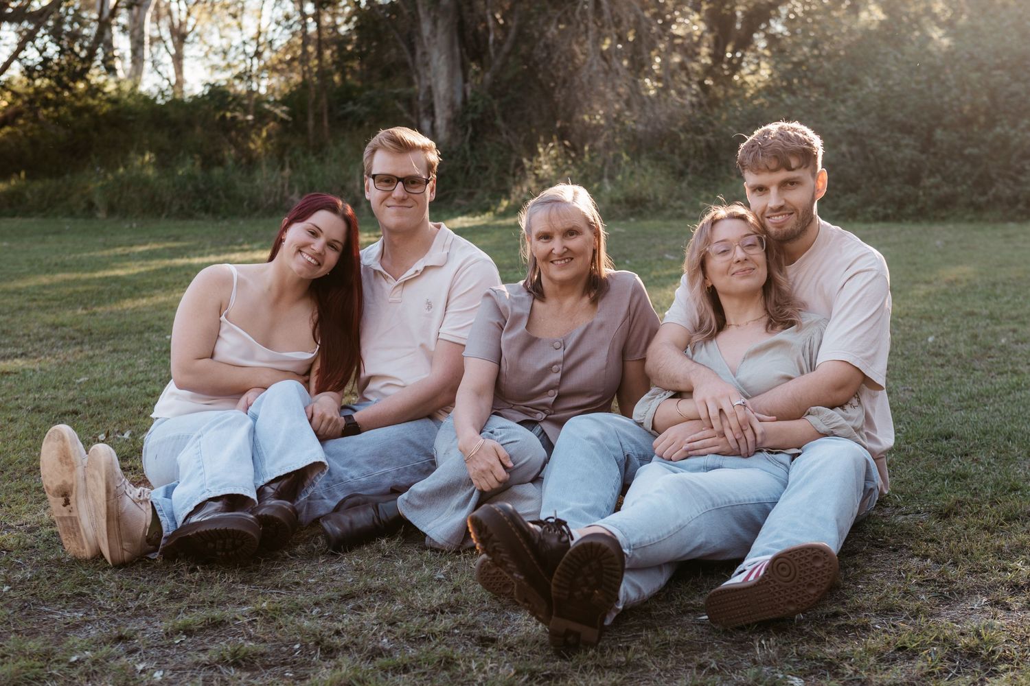 Family of five smiling and sitting together on grass outdoors during golden hour in a park setting.