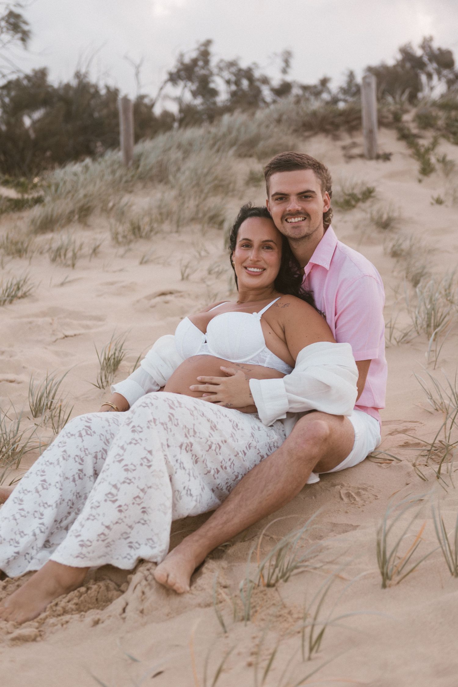 Couple sitting on sandy beach dunes, pregnant woman in white floral skirt embraced by man in pink shirt, smiling.
