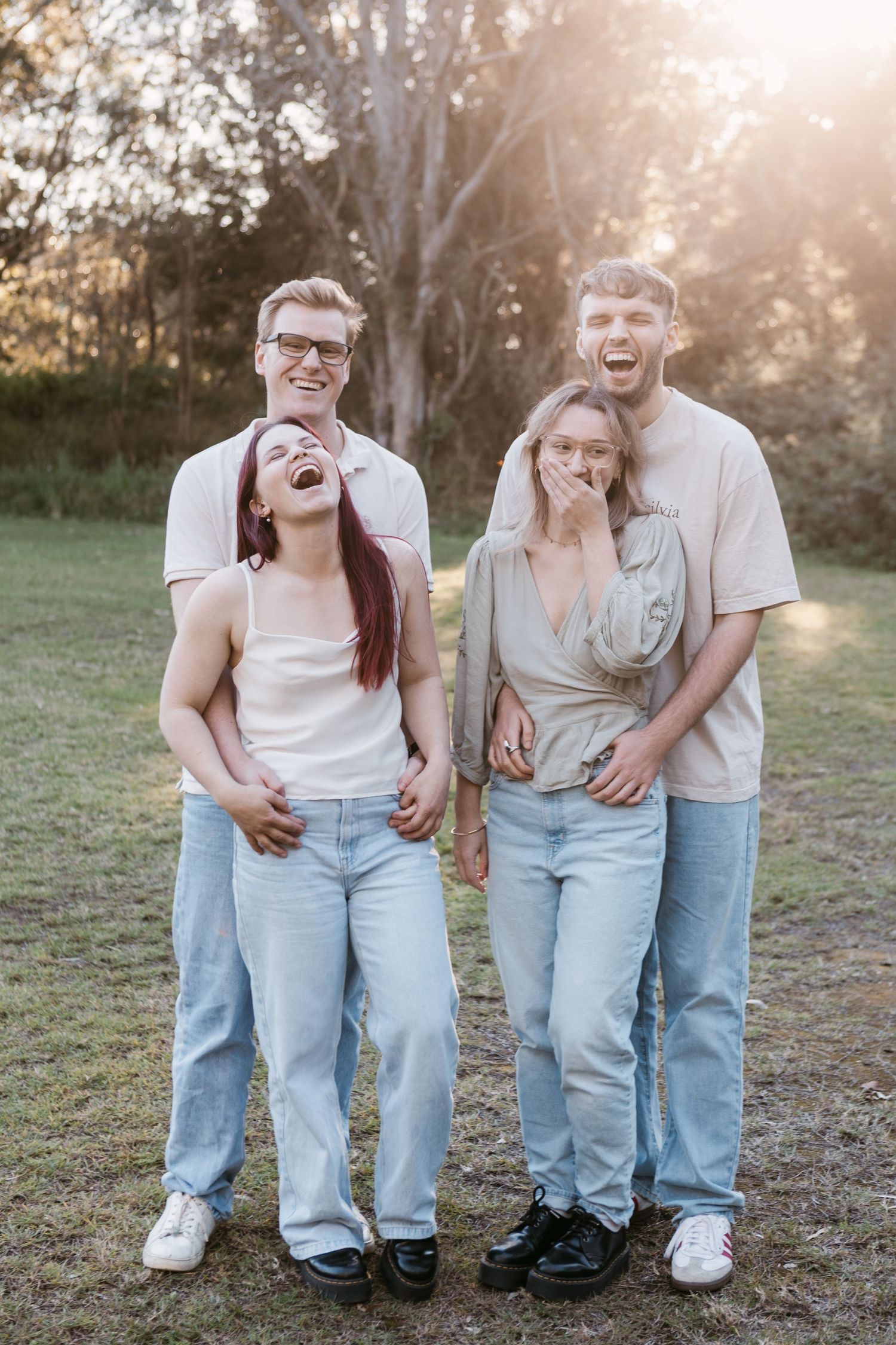 Four laughing friends pose together outdoors in casual denim outfits during golden hour in a park setting.