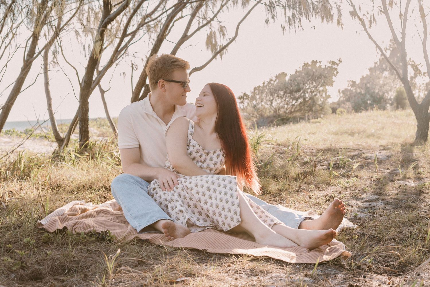 Happy couple sitting together on a blanket in a sunlit field surrounded by eucalyptus trees.