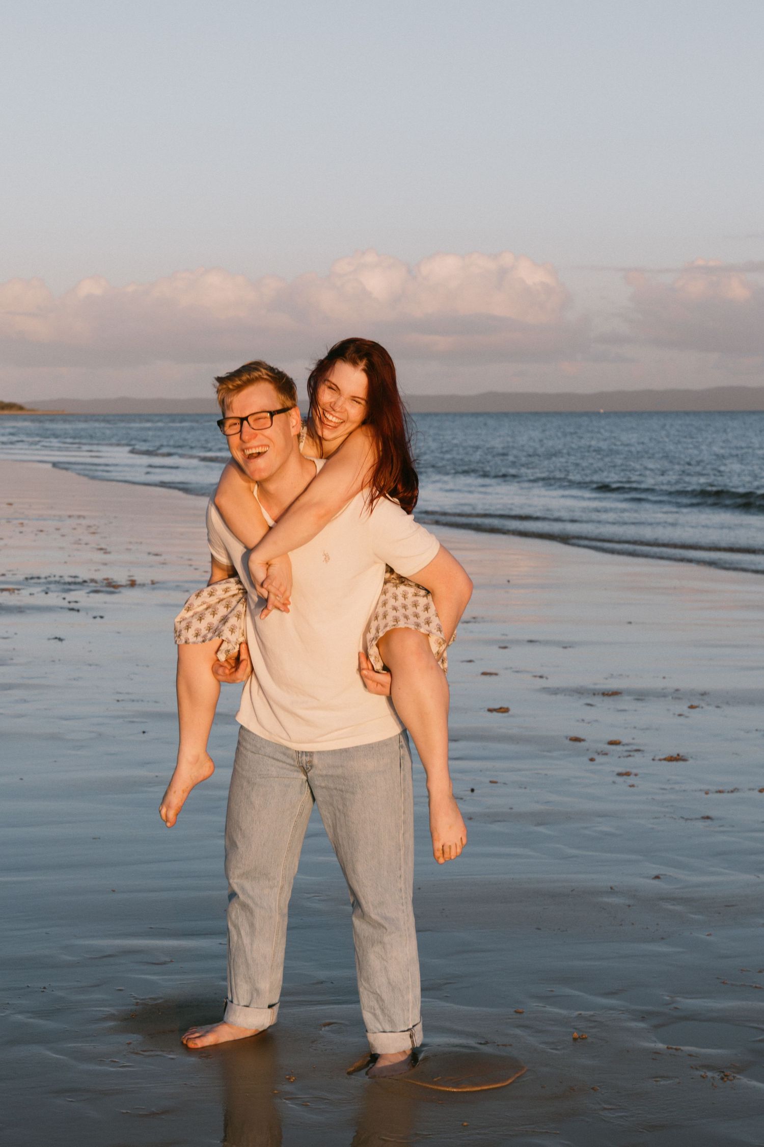 Man giving smiling woman a piggyback ride on a sandy beach at golden hour.
