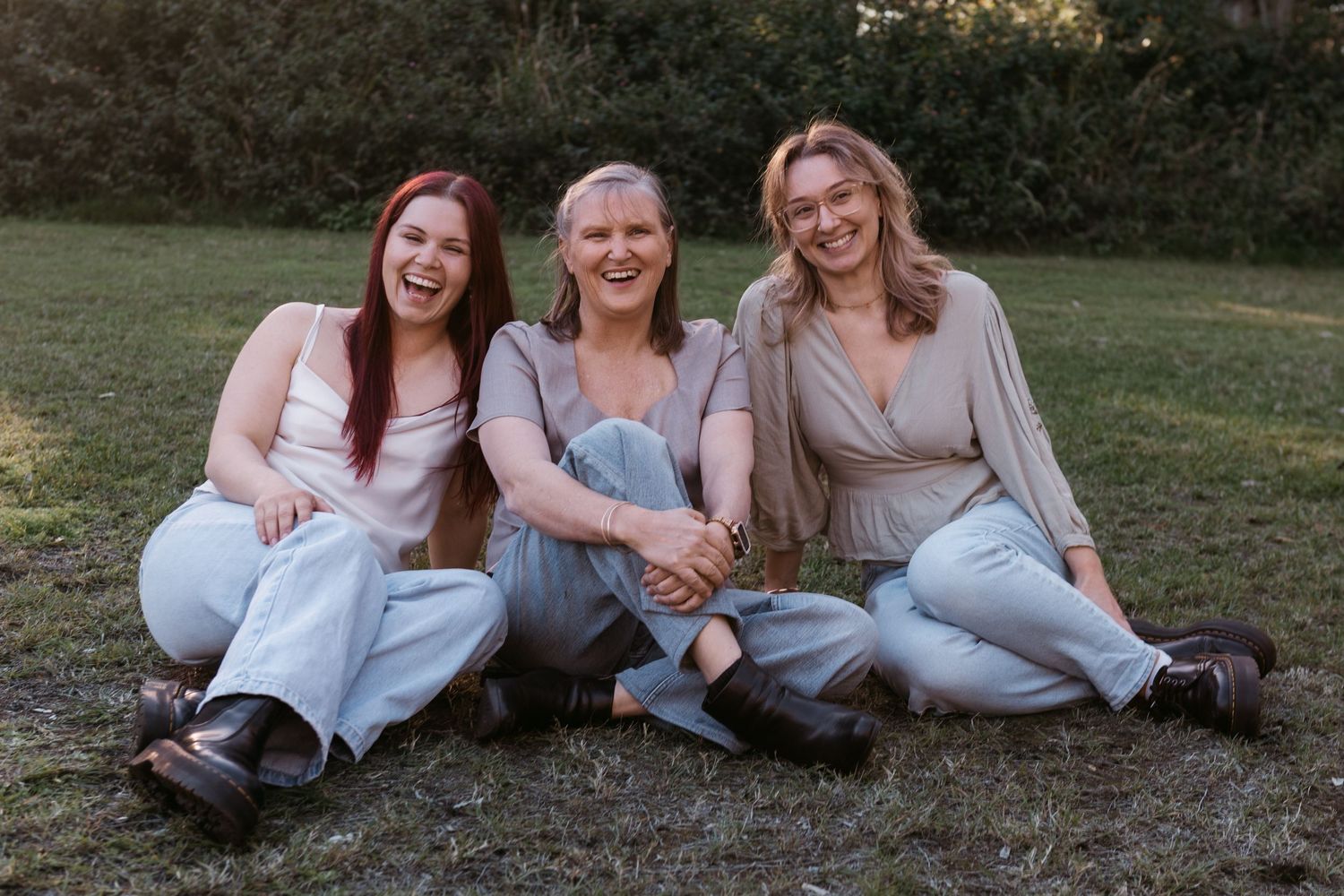 Three women sitting together on grass outdoors, smiling and laughing, wearing casual light-colored outfits.