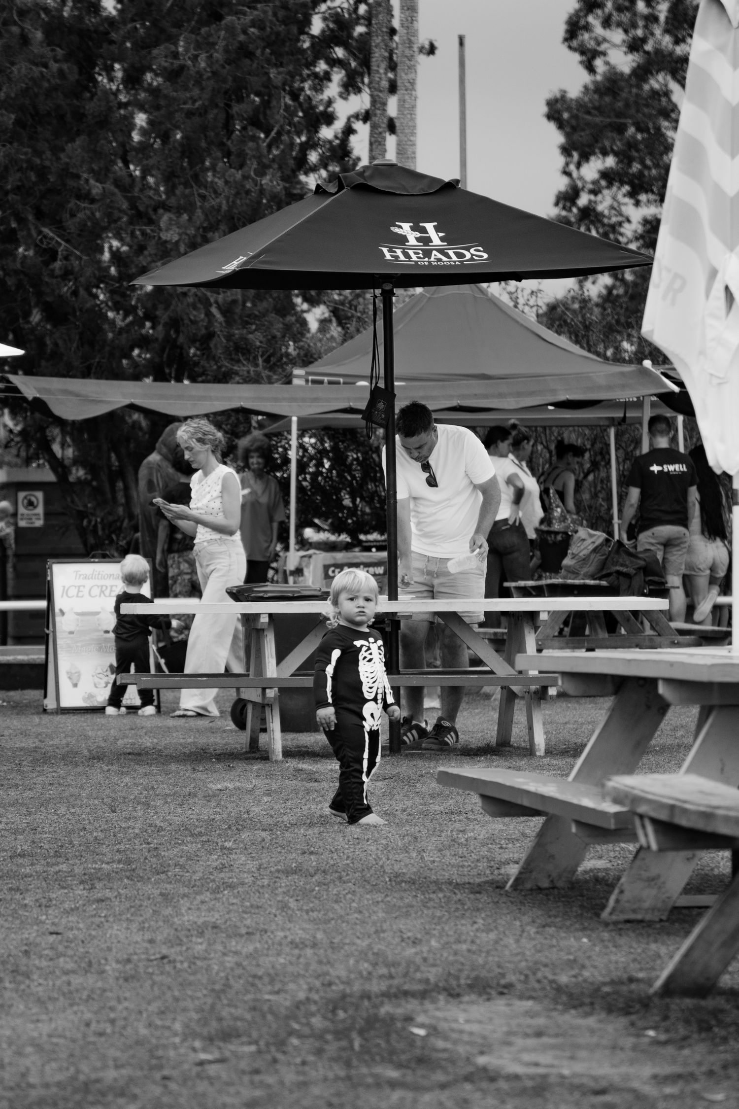 Black and white photo of a toddler in a skeleton costume walking at an outdoor Halloween event.
