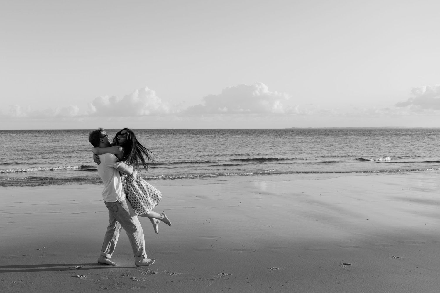 A couple kisses on a sandy beach at sunset in a romantic black and white photo.