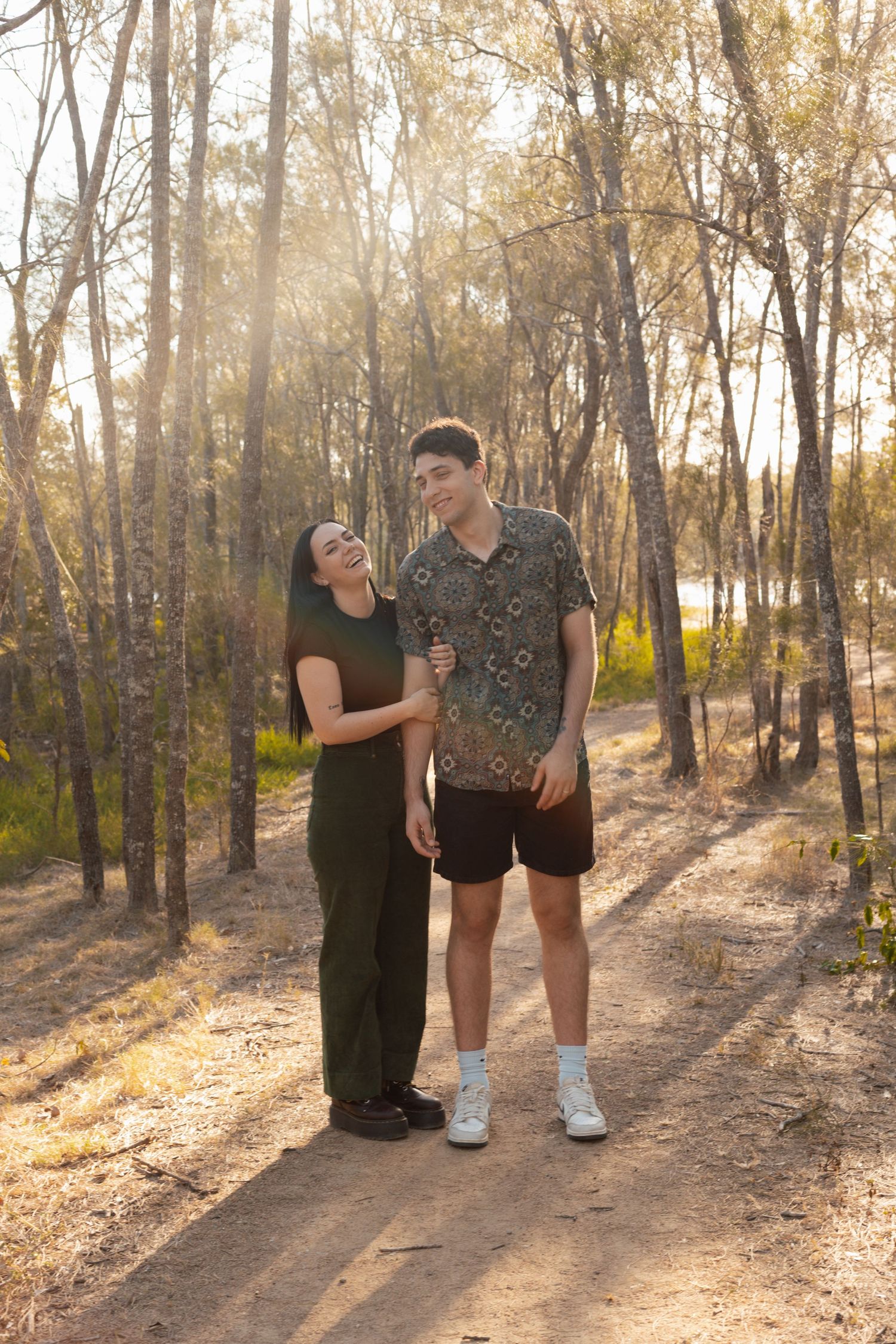 Young couple laughing together on a sunlit forest trail with tall trees in the background.