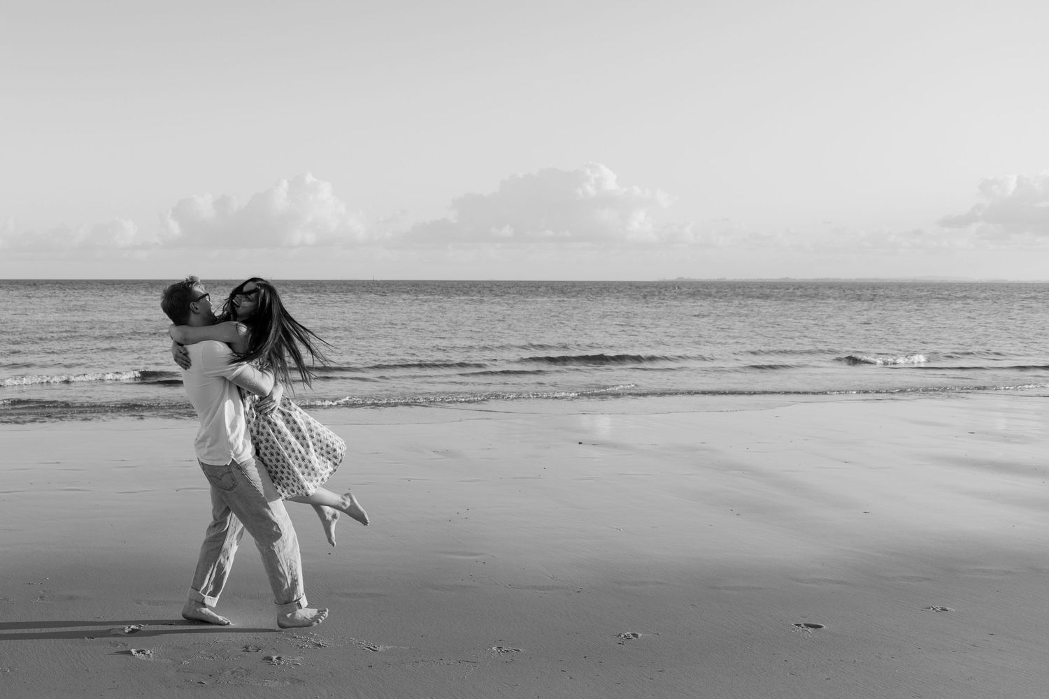Black and white romantic photo of couple embracing and kissing on a serene beach shoreline.