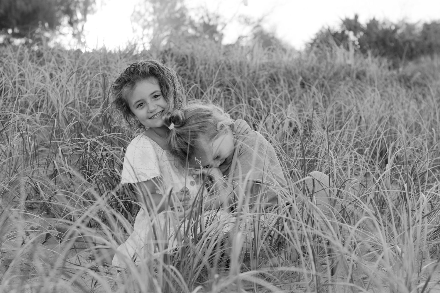 Two young children sitting together in tall grass during a family photo session.
