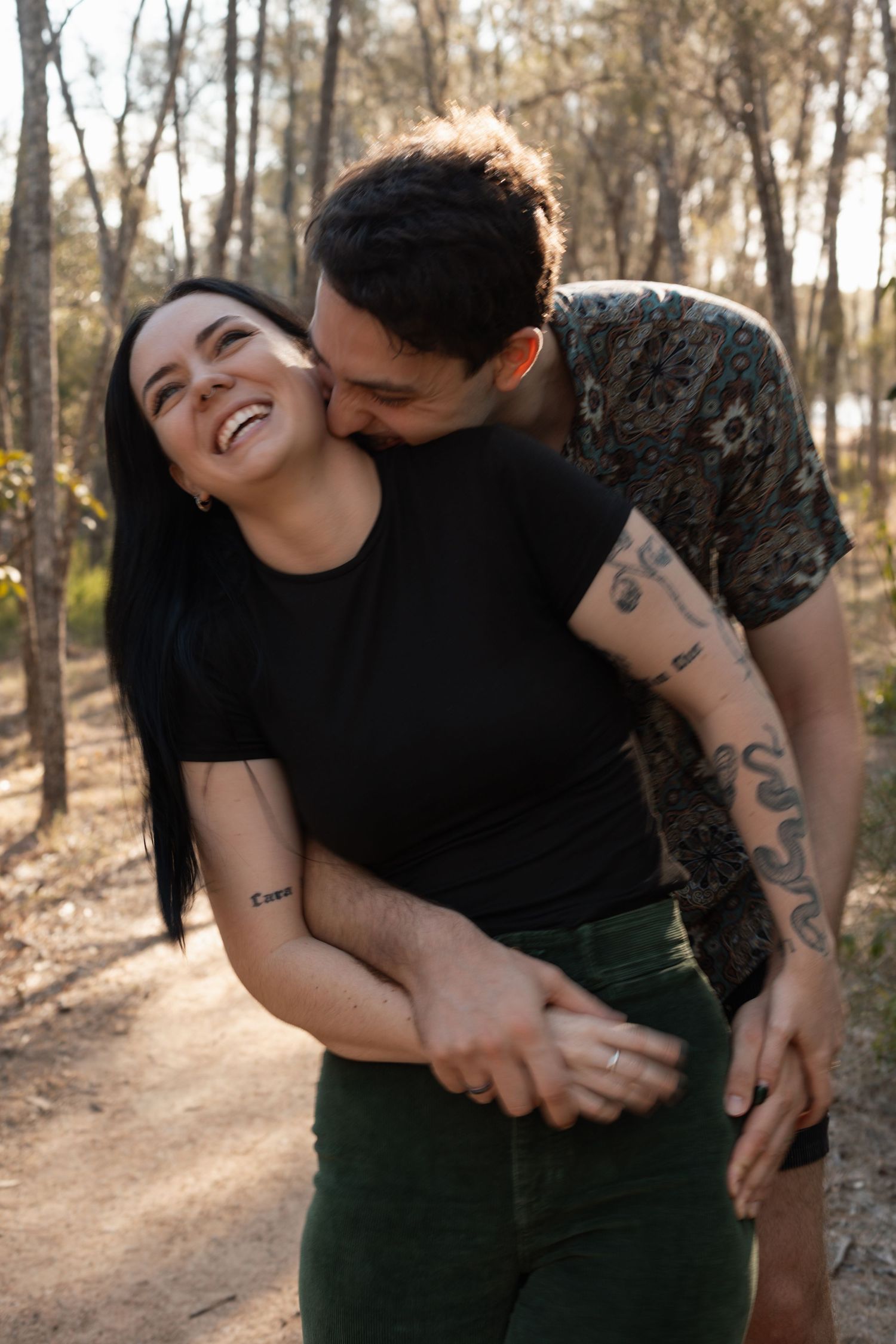 Tattooed couple embracing and laughing together on a sunlit forest path.