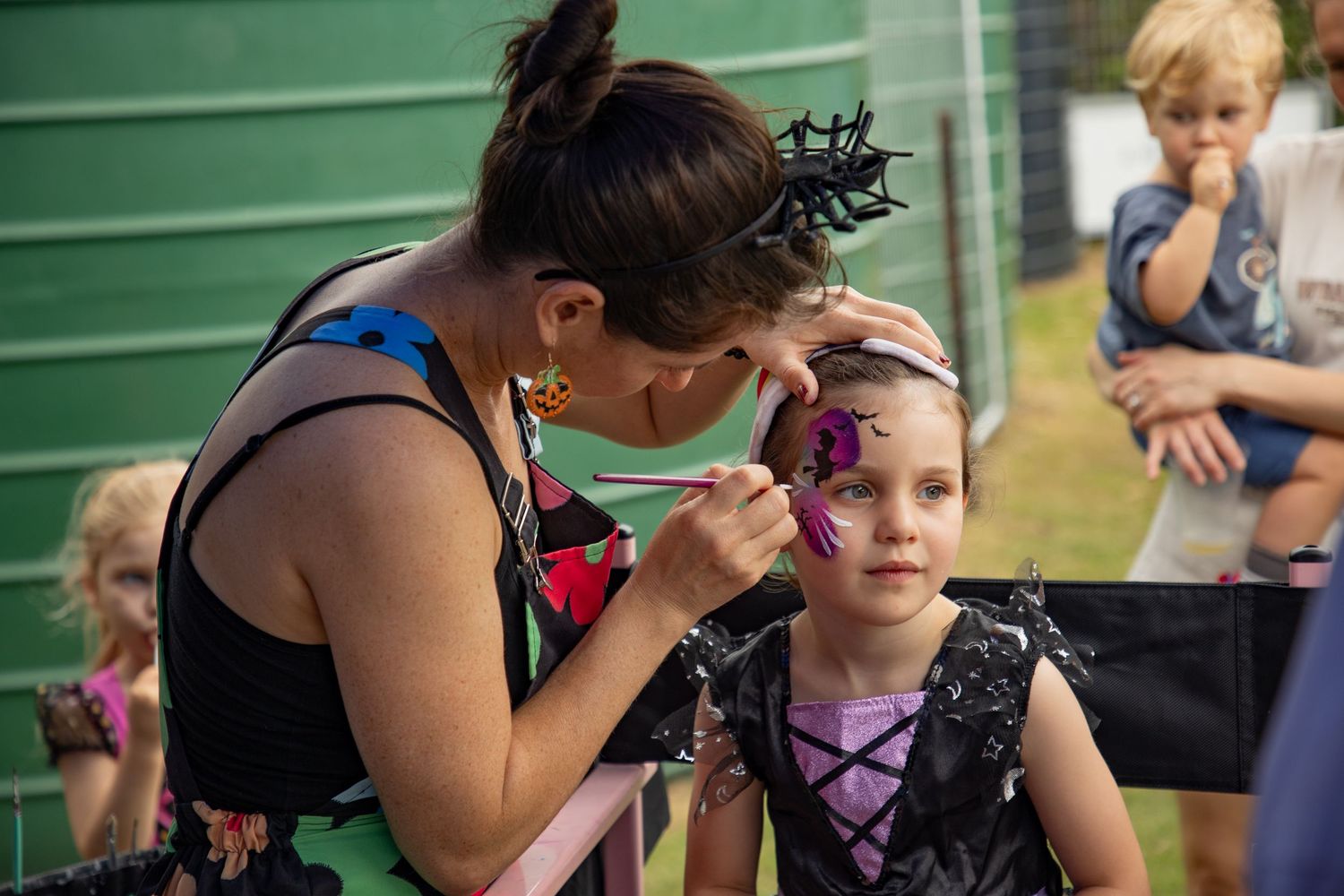 Face painter applying pink butterfly design to young girl at Halloween festival outdoor event.