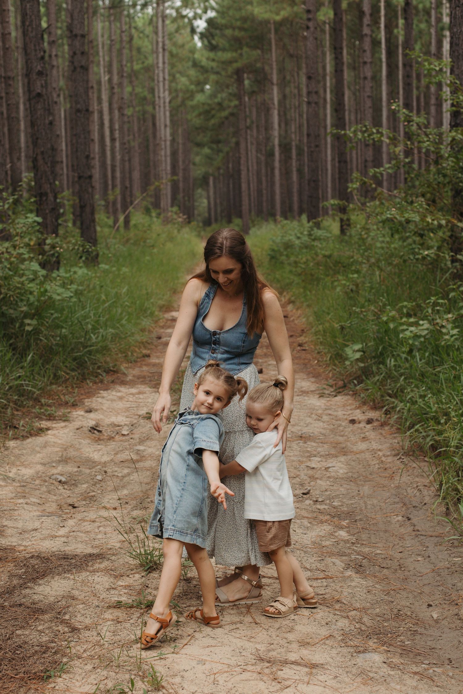 Mother bending down to embrace two young children on a pine forest dirt path.