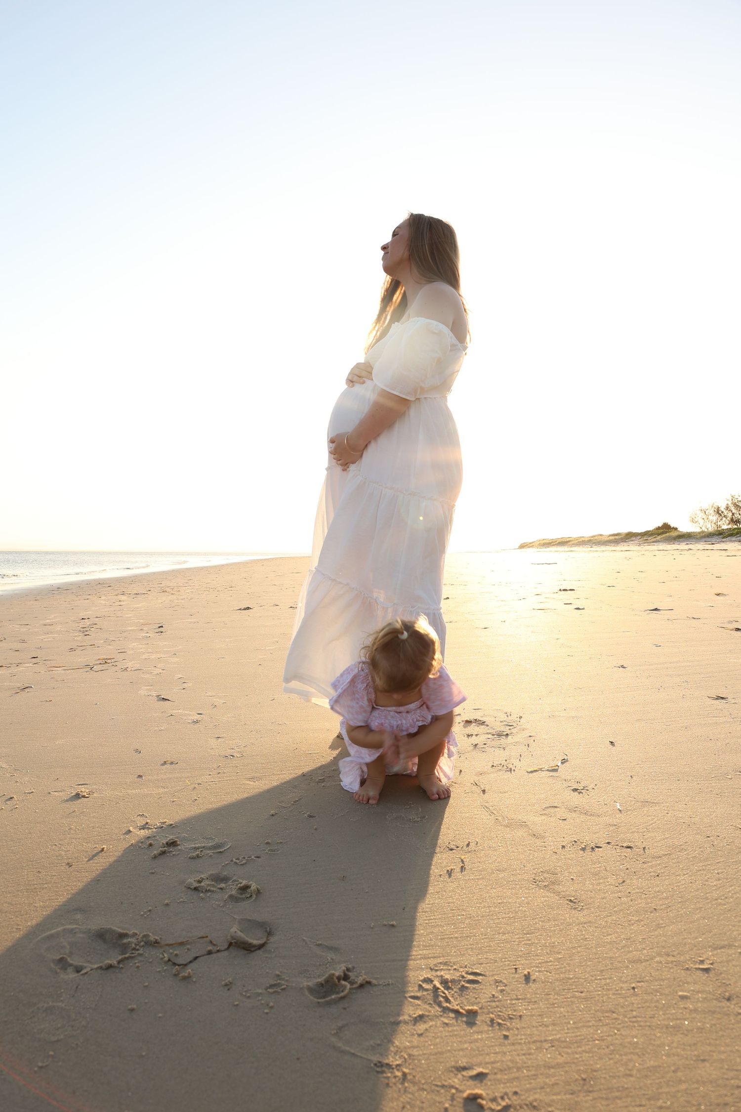 A pregnant woman in a white dress stands on a sunny beach as a toddler plays in the sand.