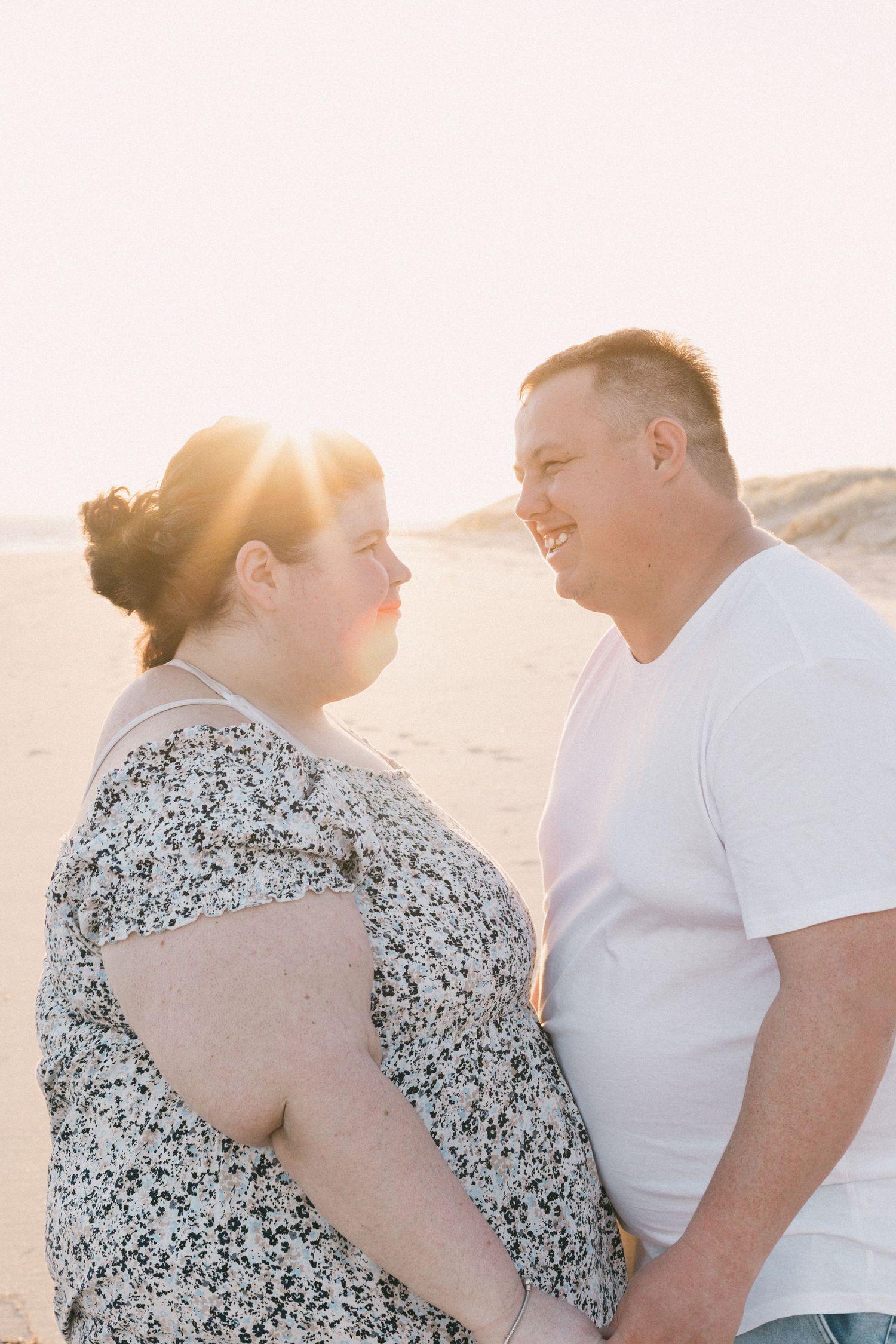 Happy couple smiling at each other on sandy beach with golden sunset backlight behind them.