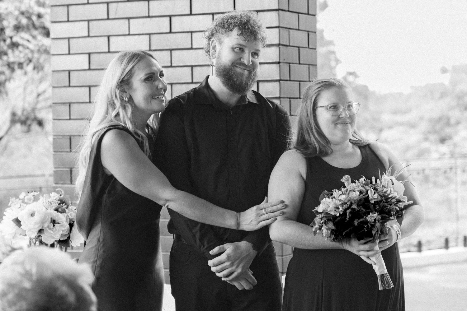 Two bridesmaids in black dresses flanking groomsman holding bouquet at outdoor wedding ceremony.
