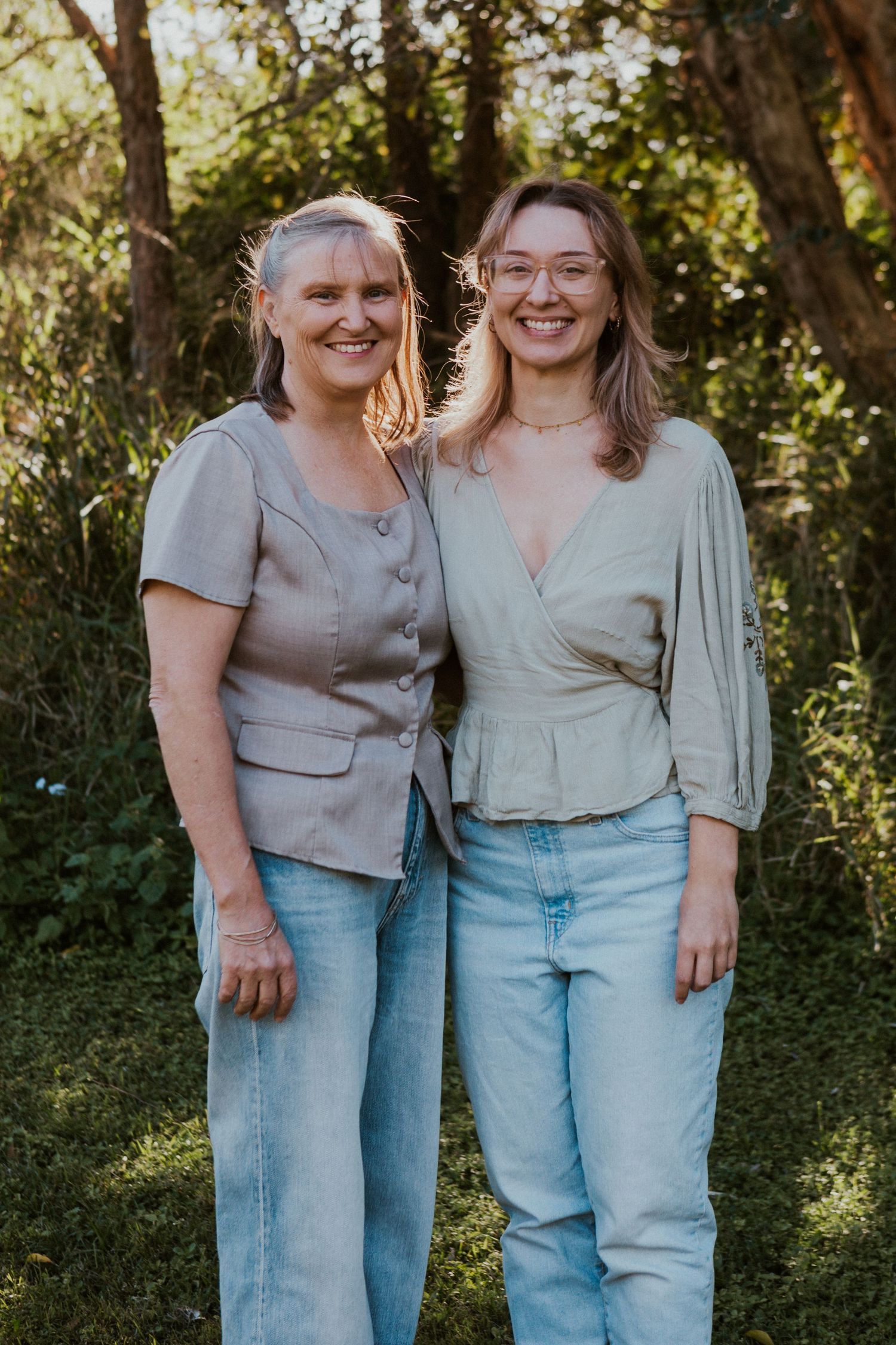 Two smiling women in casual sage green tops and jeans pose together outdoors in a lush green setting.