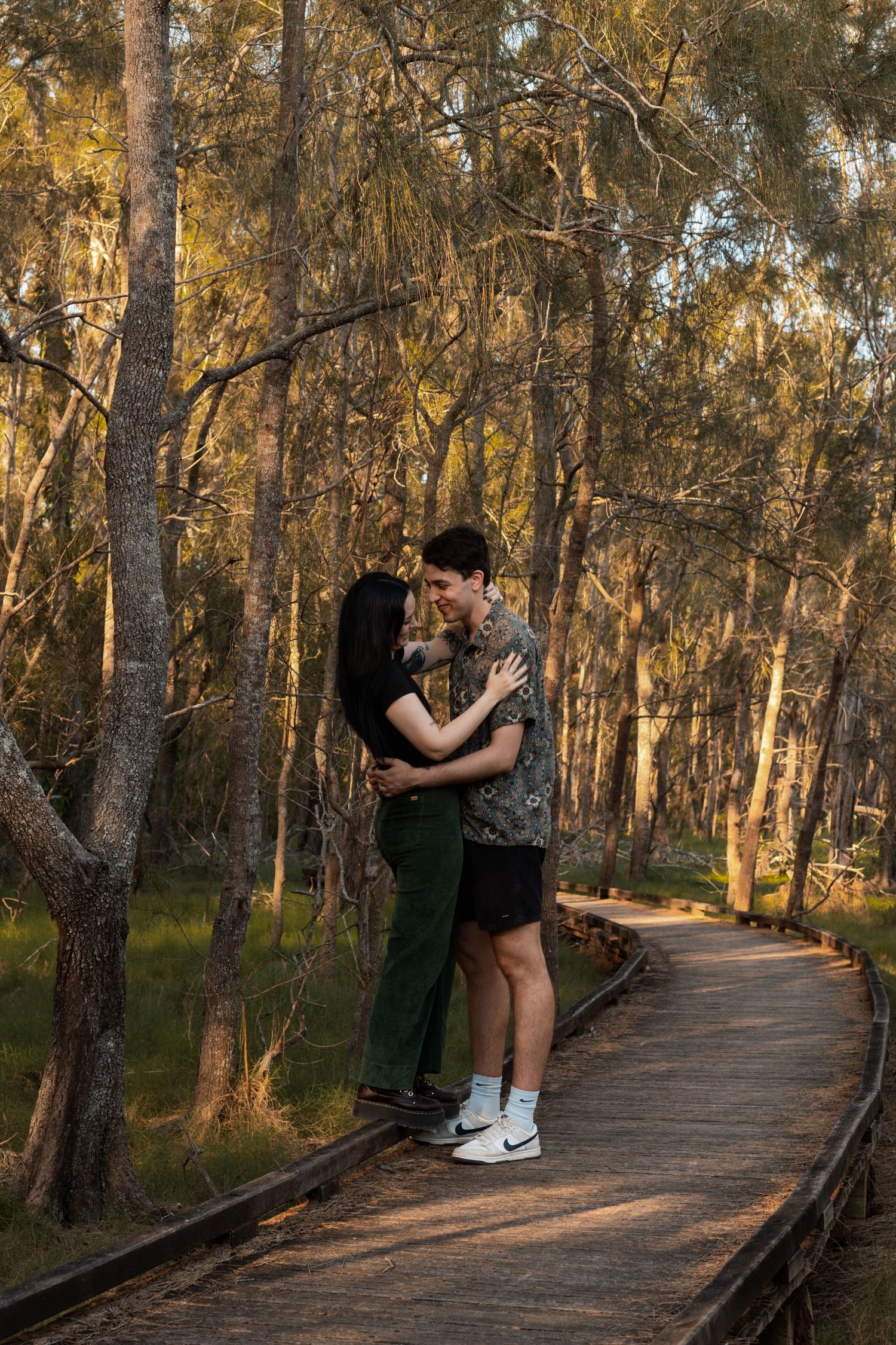 A couple embracing and smiling on a wooden boardwalk surrounded by golden sunlit forest trees.