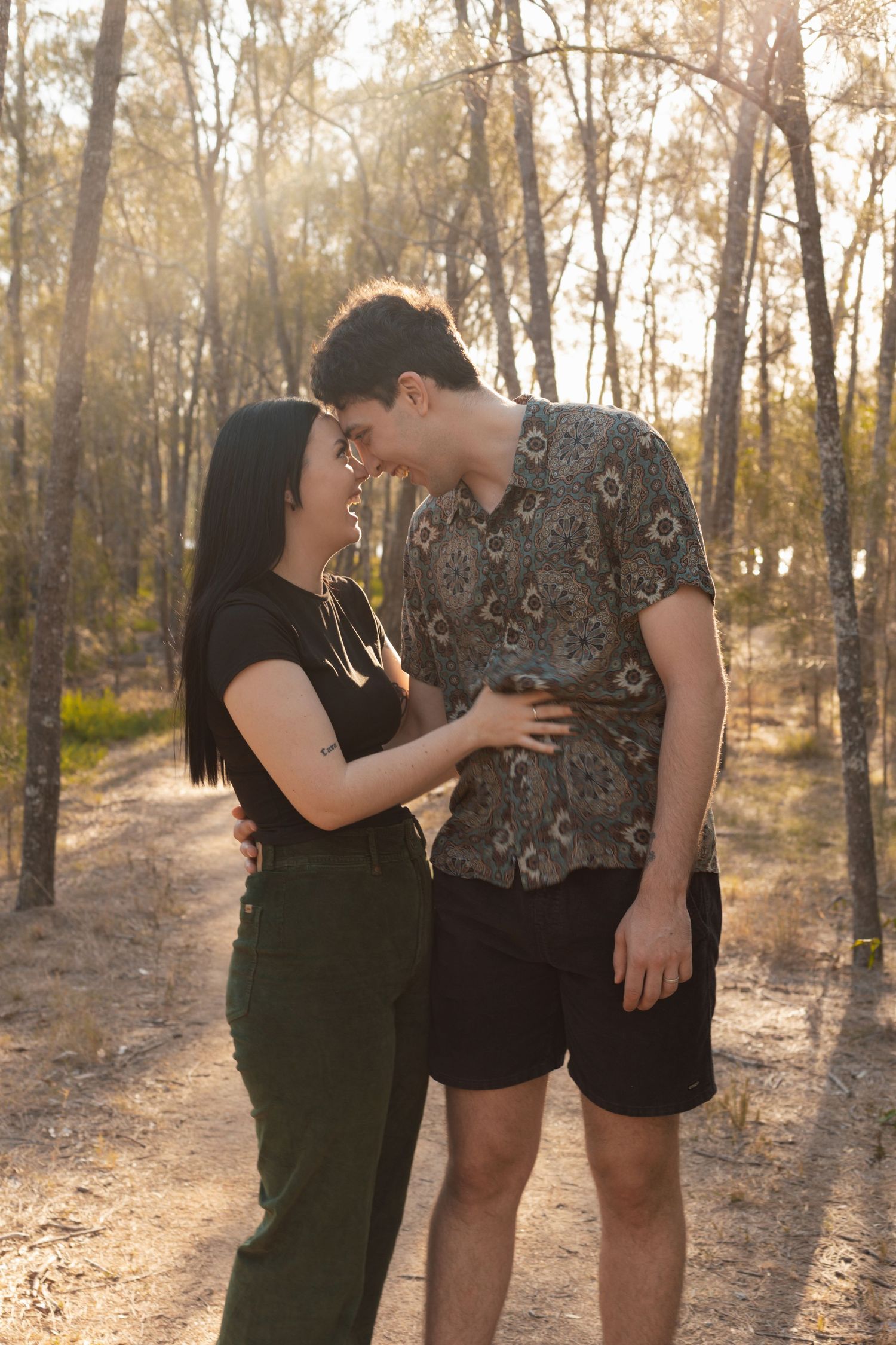 Couple sharing an intimate nose-to-nose moment on a sun-drenched forest path surrounded by tall trees.