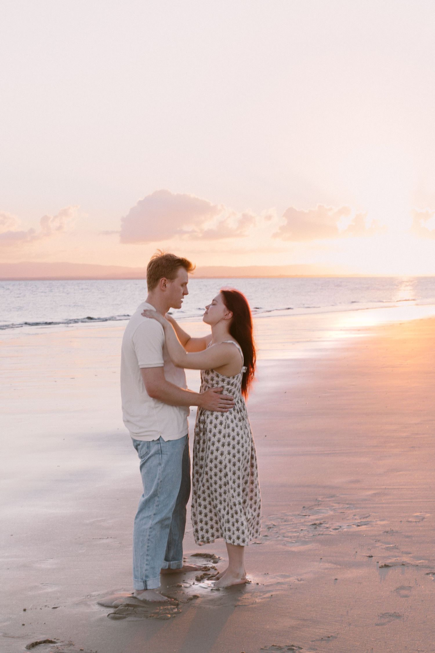 Romantic couple embracing on a beach at golden sunset, woman in floral sundress.