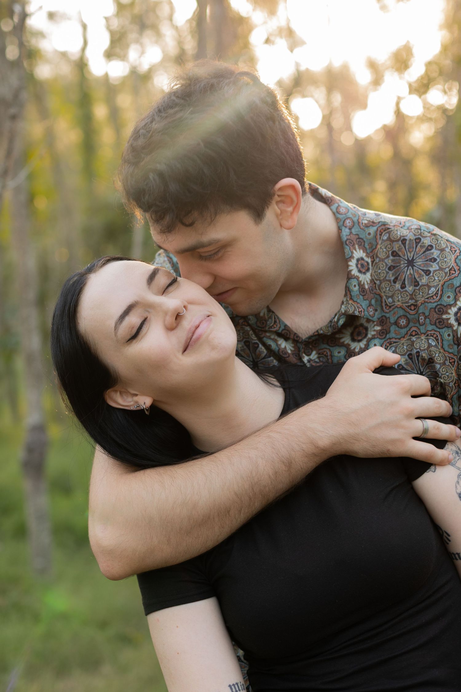 Couple sharing a tender moment in a sunlit forest, man kissing womans cheek while embracing her.