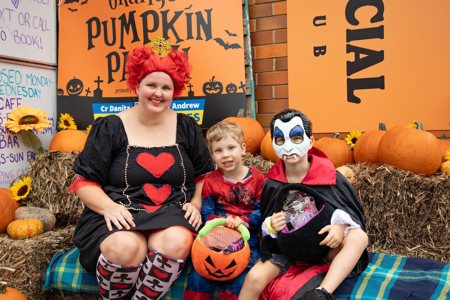 Woman and two children in Halloween costumes pose at a pumpkin patch display with hay bales and pumpkins.