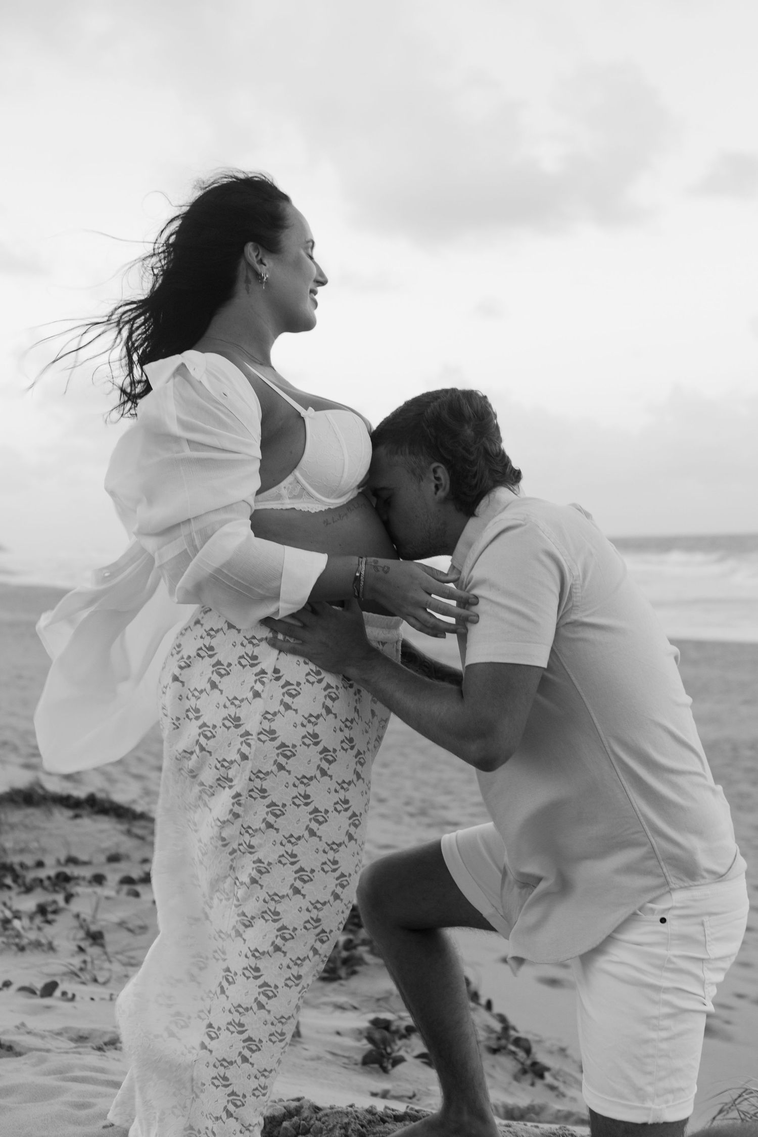 Black and white photo of a man kissing pregnant woman's belly on a beach, her hair blowing in the wind.